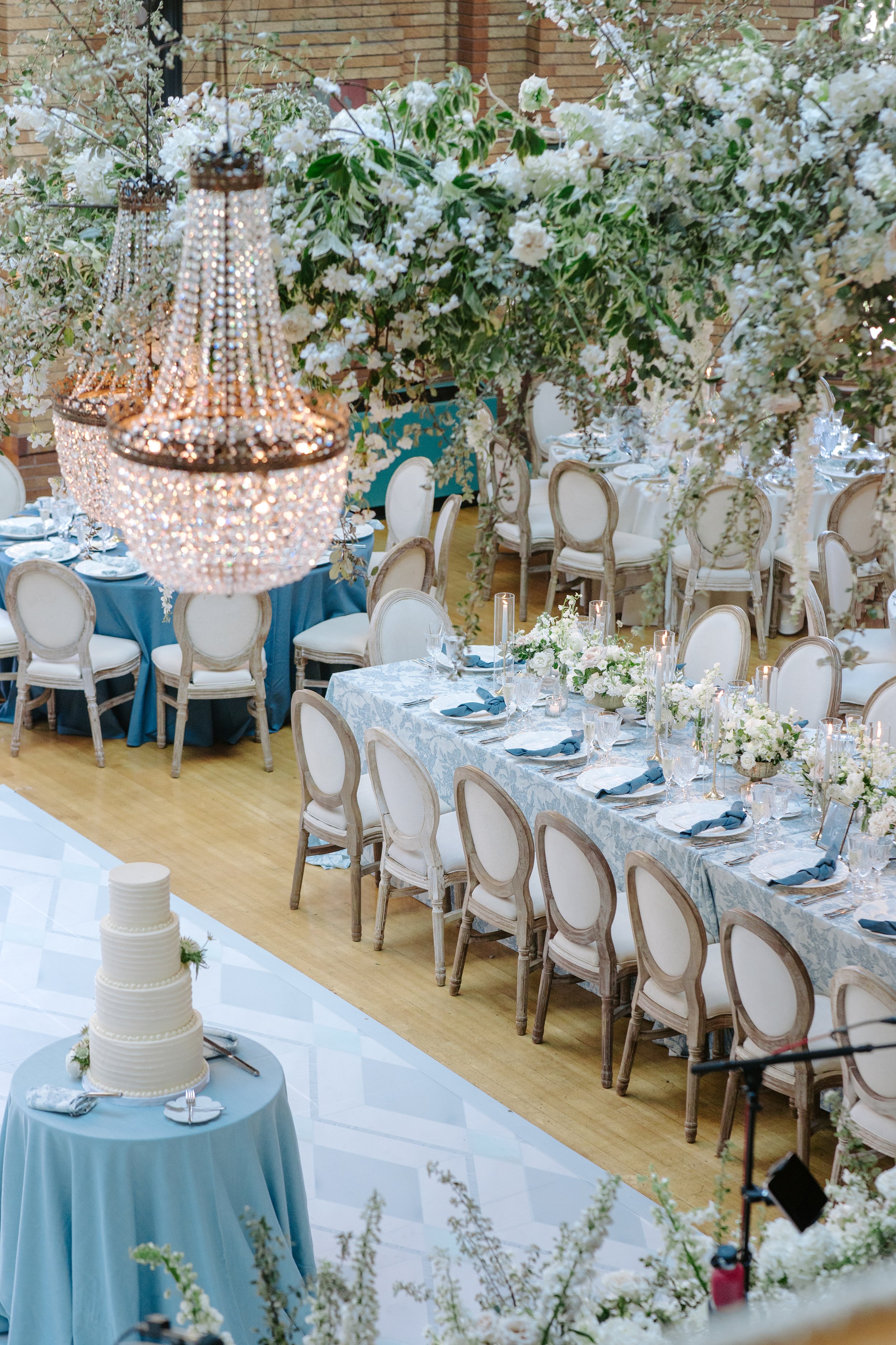 Elegant wedding reception with long tables decorated with white floral centerpieces, surrounded by white chairs with wooden frames, and a tall multi-tiered white wedding cake on a separate table. The scene is enhanced by romantic lighting and a large
