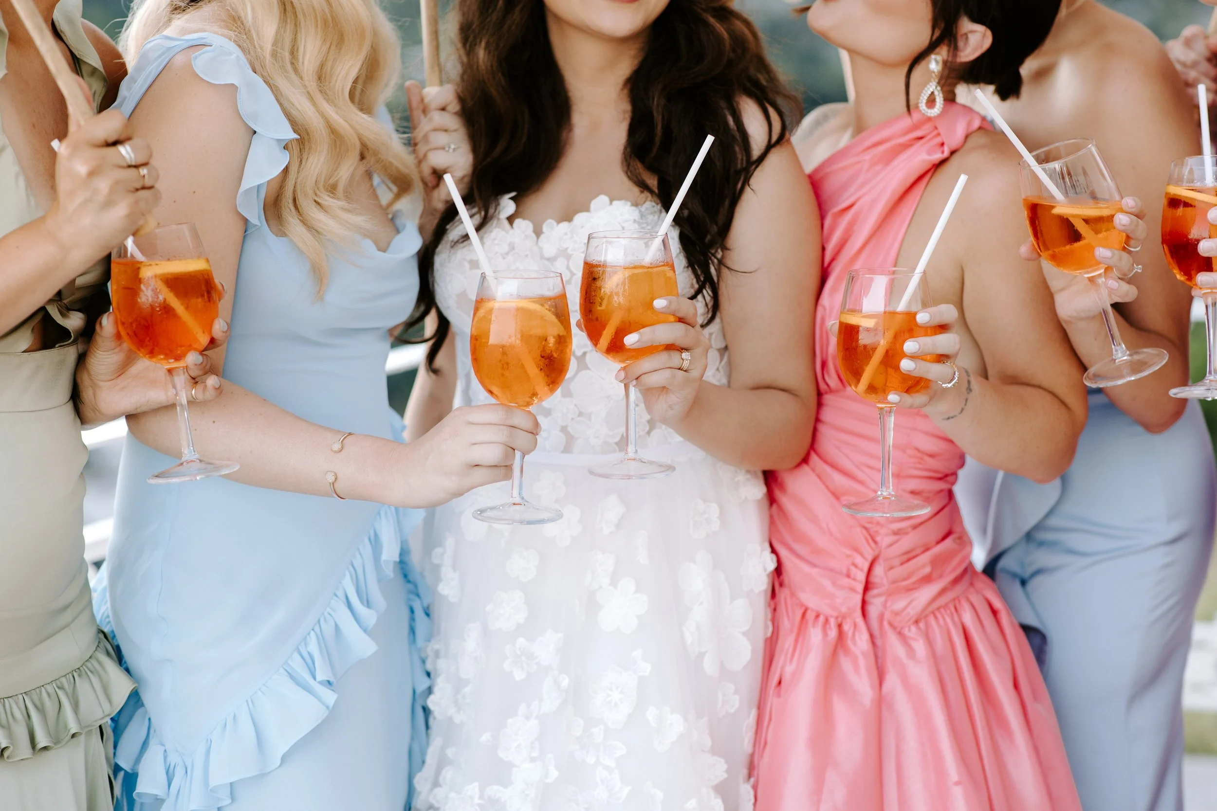 Group of women in colorful dresses holding glasses of orange-colored drink with ice and straws at an outdoor gathering.