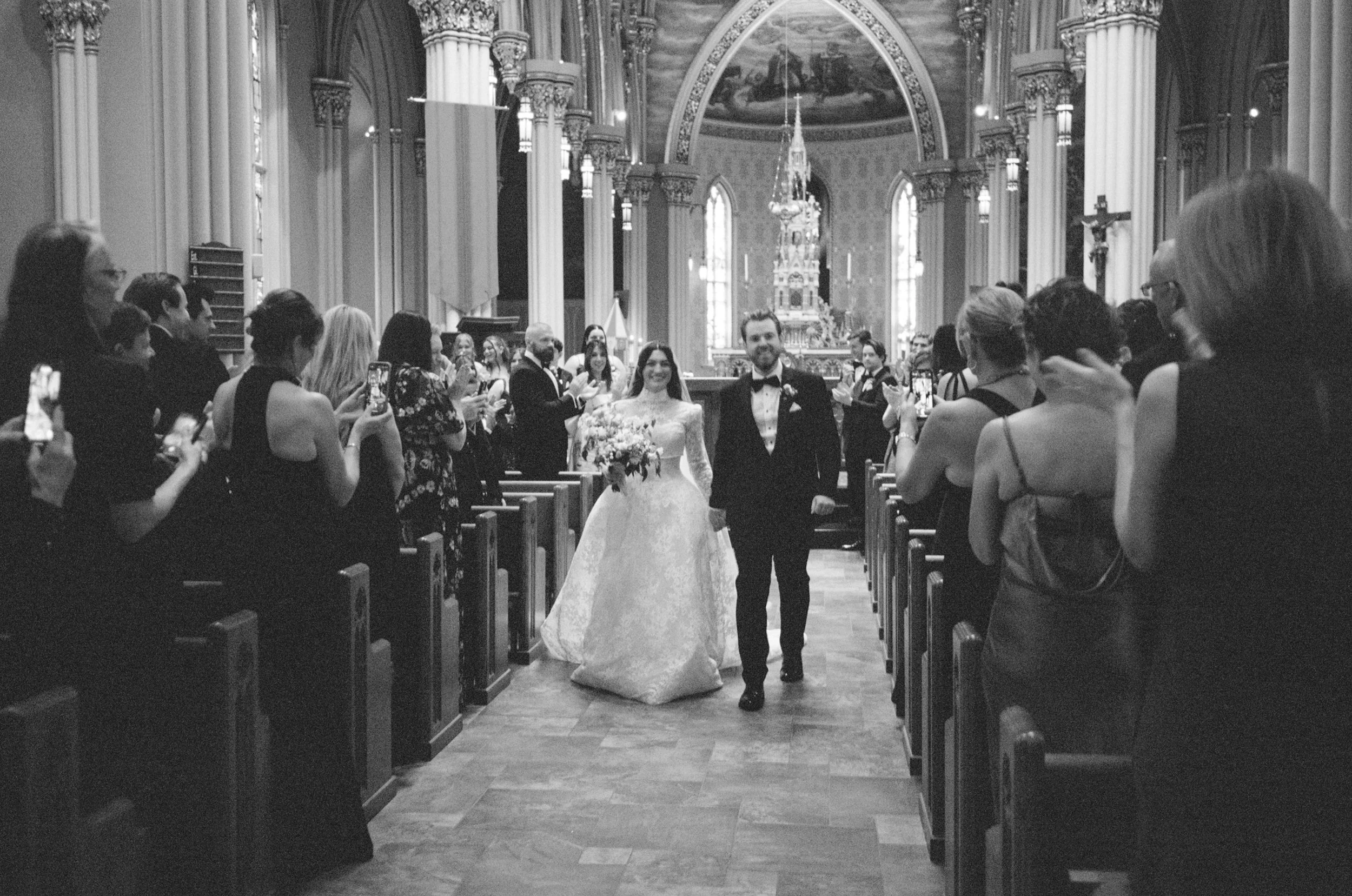 A bride and groom walking down the aisle of a church, smiling, surrounded by guests taking photos. The church has tall columns and an ornate altar.