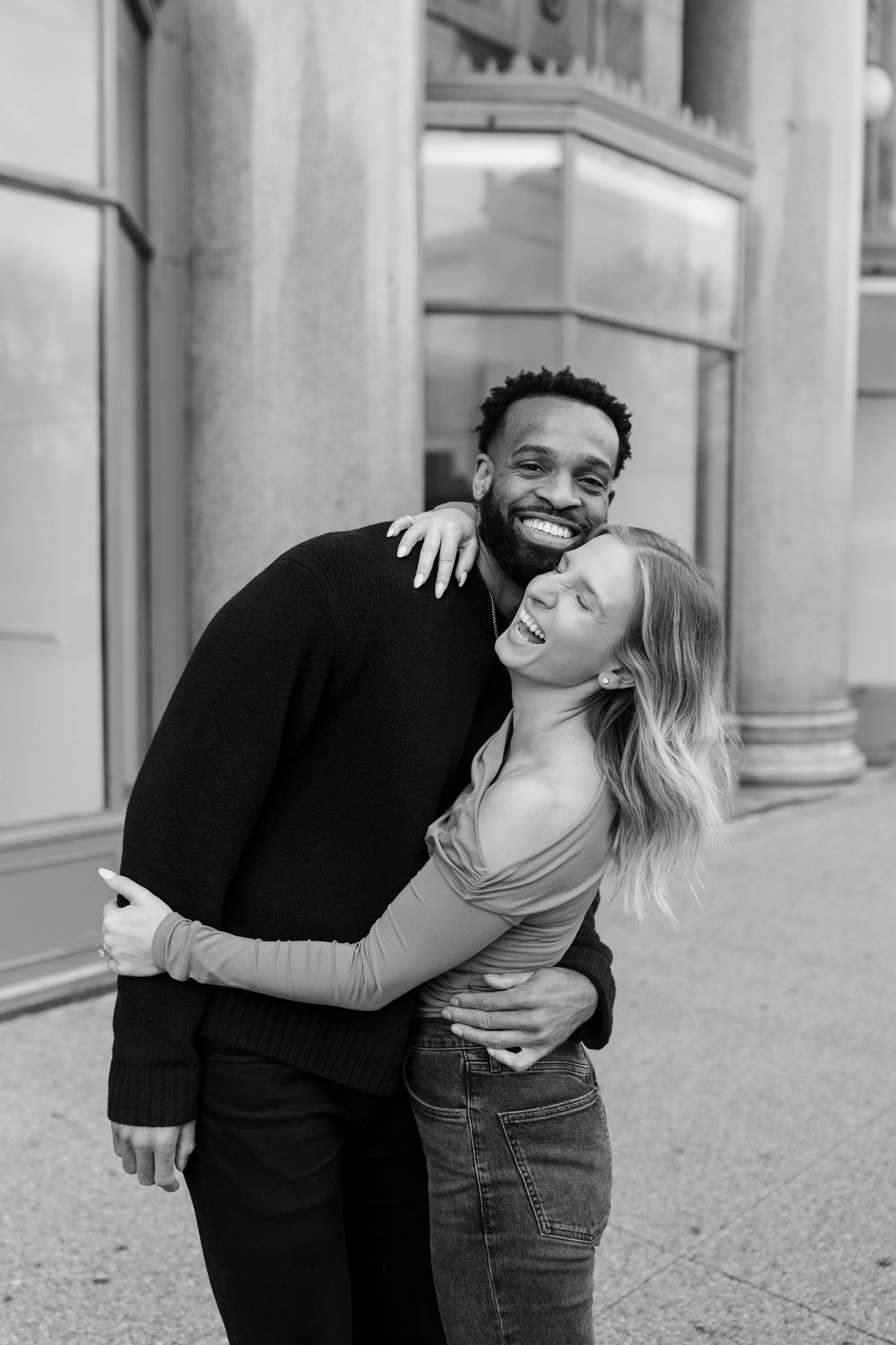 A black-and-white photo of a smiling man and woman hugging outdoors in front of a building. The man has dark skin, curly hair, and a beard, wearing a dark sweater. The woman has light skin, long wavy hair, and is wearing a long-sleeve top.