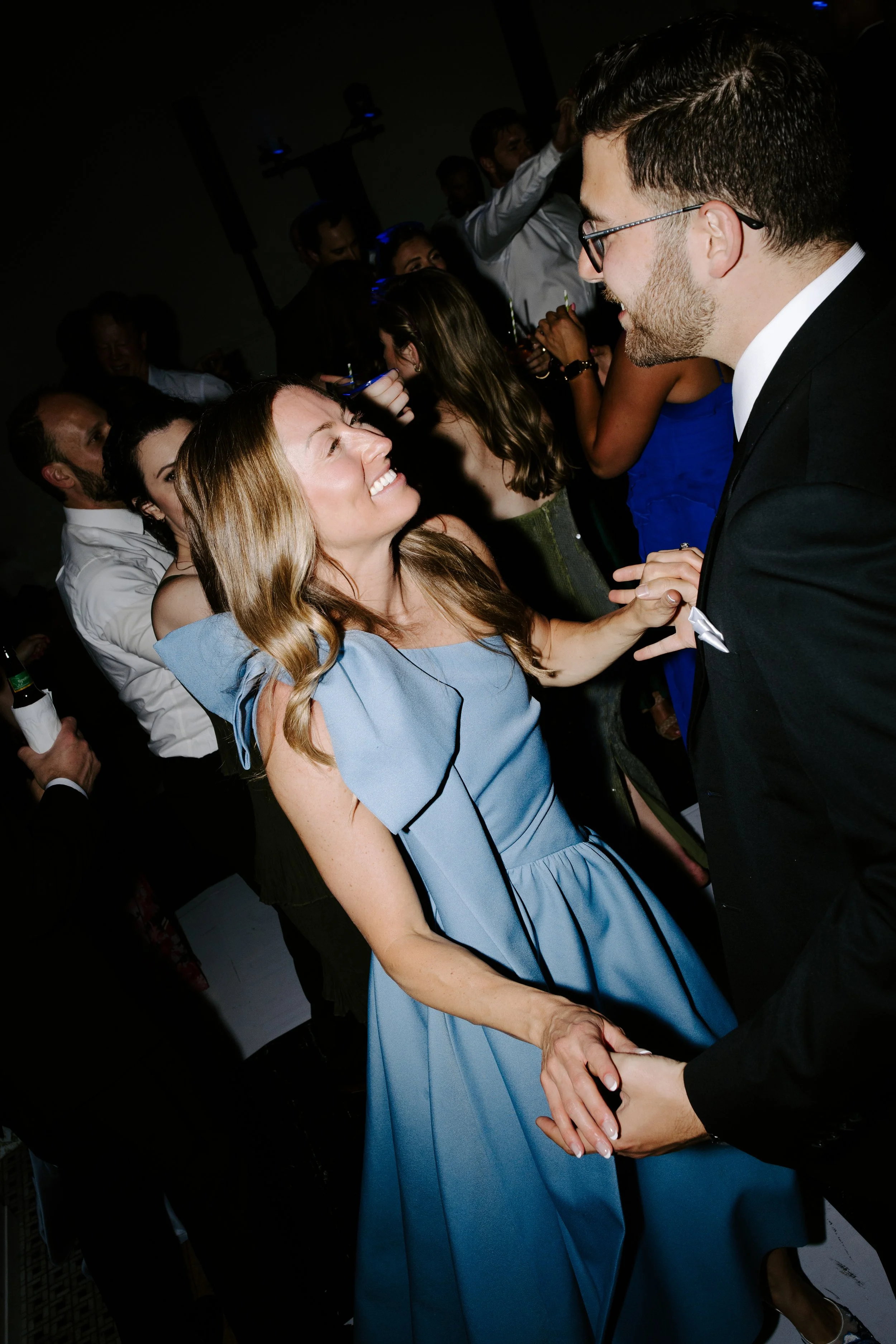 A couple dancing closely at a social event, with the woman smiling and the man wearing glasses and a tuxedo. Other people are visible in the background, some holding drinks, in a dimly lit venue.