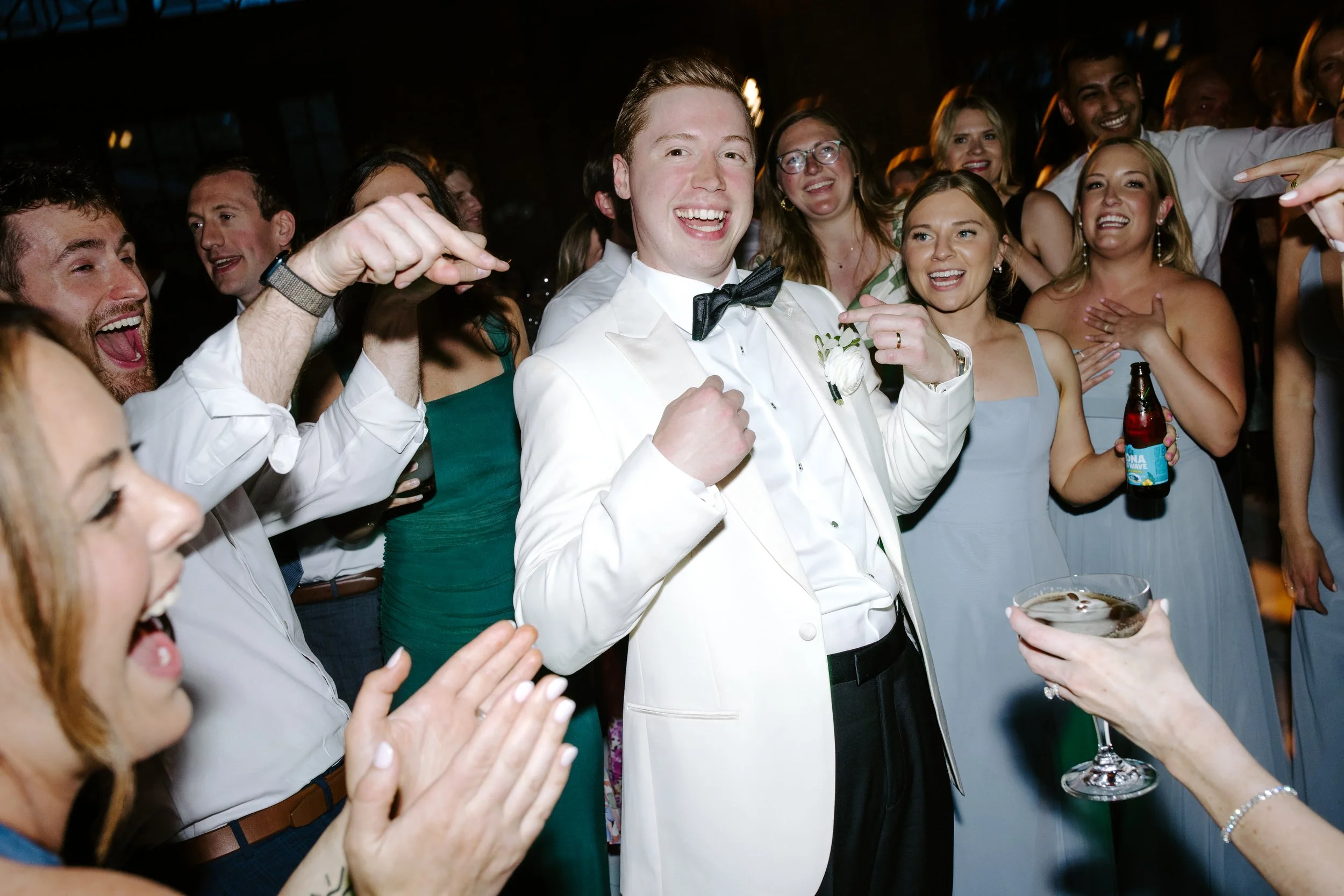 A group of people celebrating at a party, centered around a smiling man in a white tuxedo with a black bow tie. The group appears joyful, with many clapping and smiling, and some holding drinks.