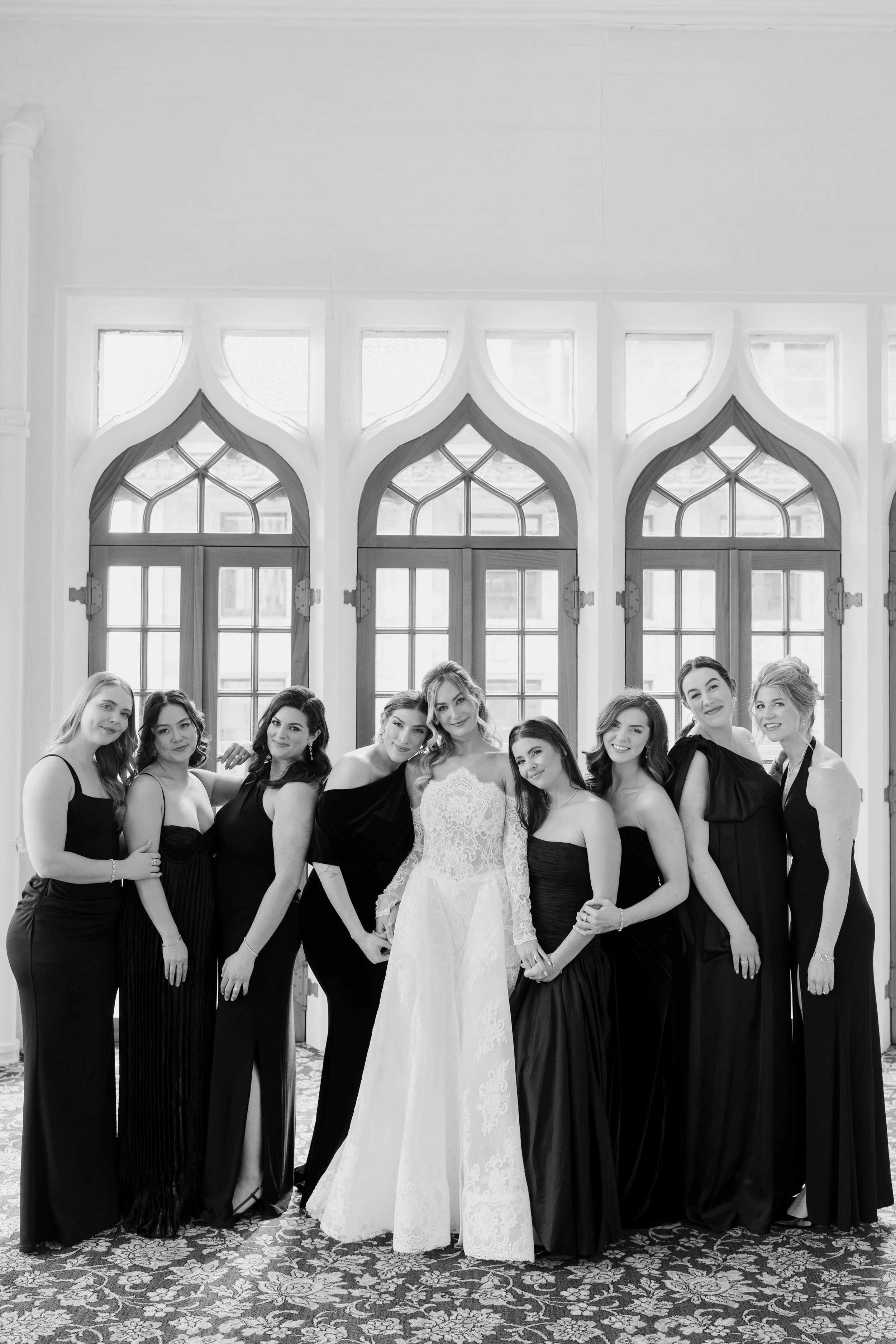 Black and white photo of a group of women dressed in formal gowns, posing indoors in front of large decorative windows, with floral carpet on the floor.