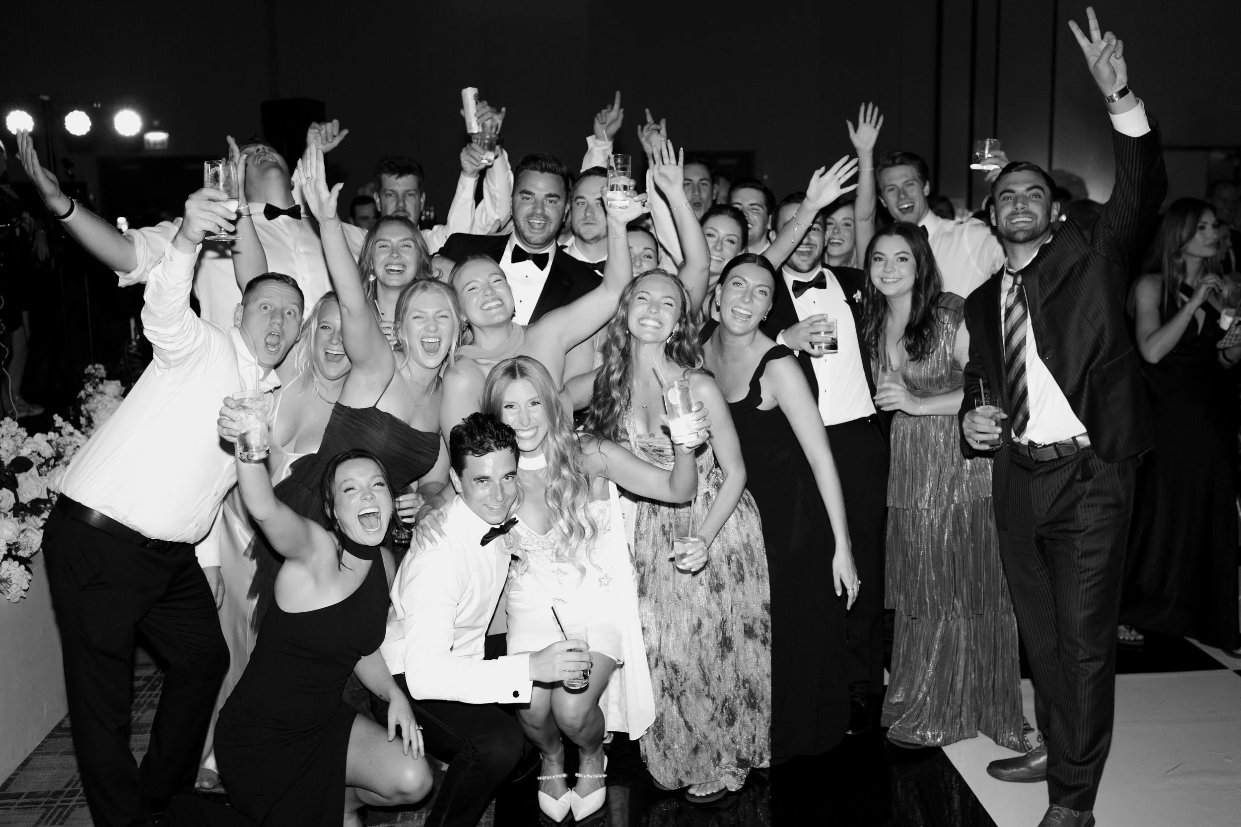 A group of people dressed in formal attire at a celebration, smiling and holding drinks, with some raising their hands and making peace signs.