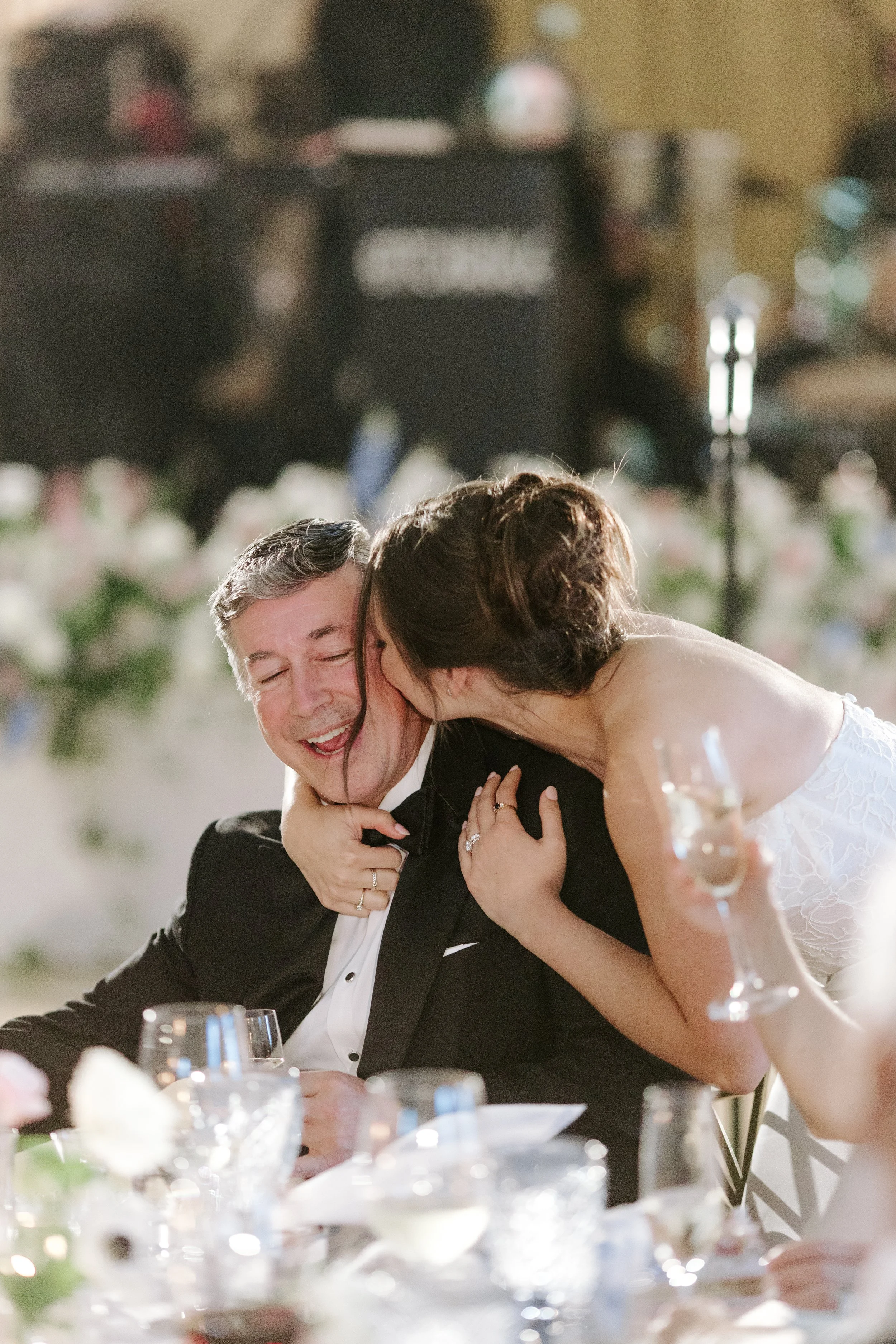 A bride and groom sharing a joyful moment at their wedding reception, with the bride leaning in to kiss the groom on the cheek.