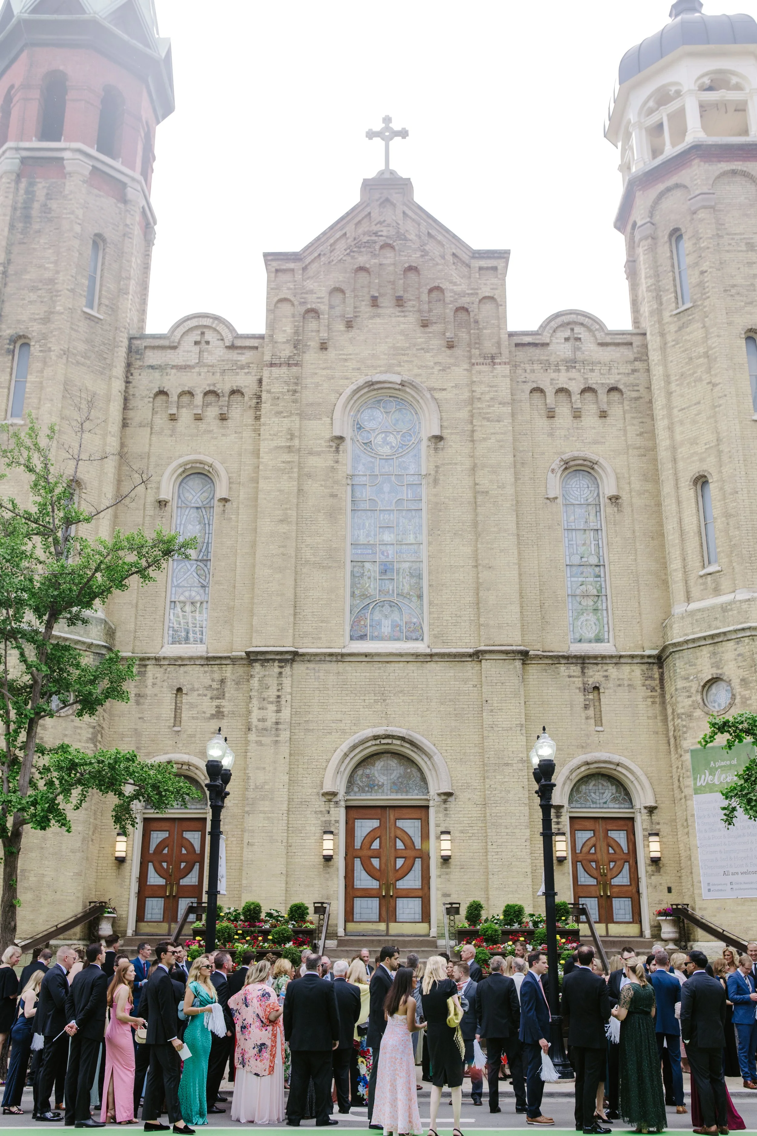 A large crowd of people dressed in formal attire gathered in front of a church with stained glass windows and twin towers, during an outdoor event.