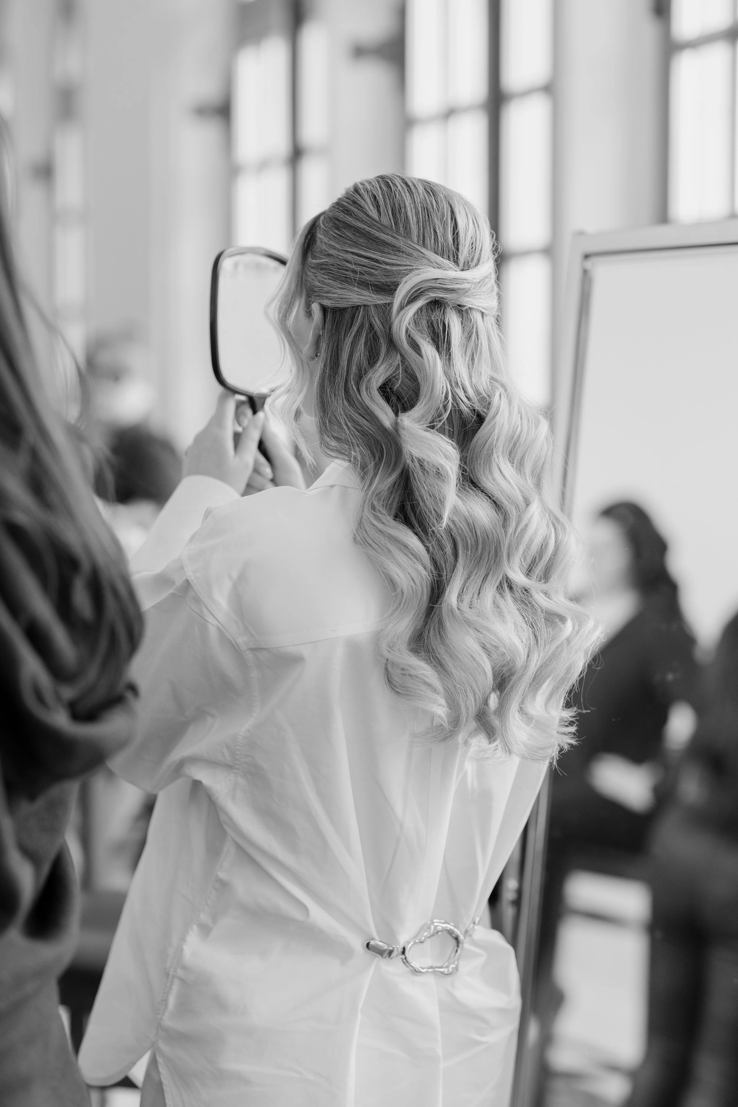 A woman with long, wavy blonde hair, dressed in a white lab coat, holding a handheld mirror and looking into it in a bright, modern room with large windows.