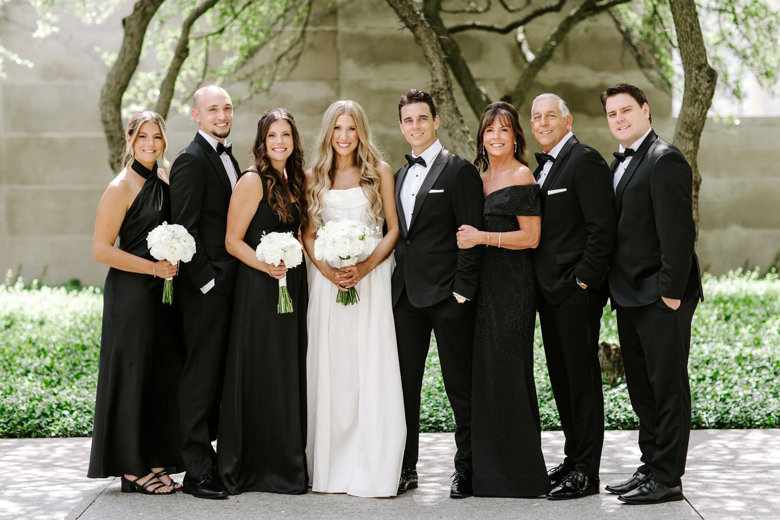 Group of eight people in formal attire posing outdoors in front of trees, with two women holding bouquets, at a wedding or formal event.