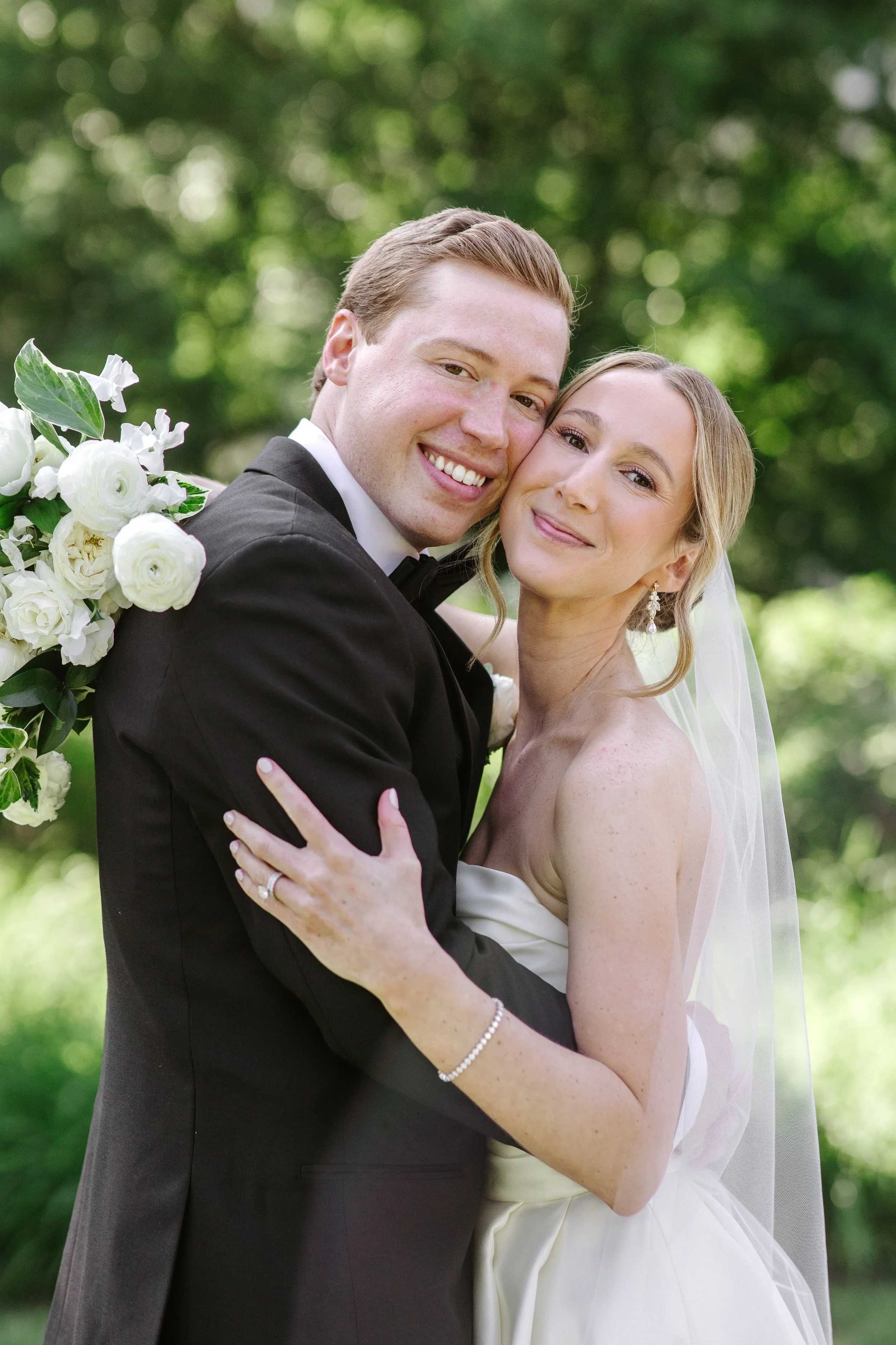 A happy newlywed couple, dressed in wedding attire, embracing outdoors with green blurred trees in the background.