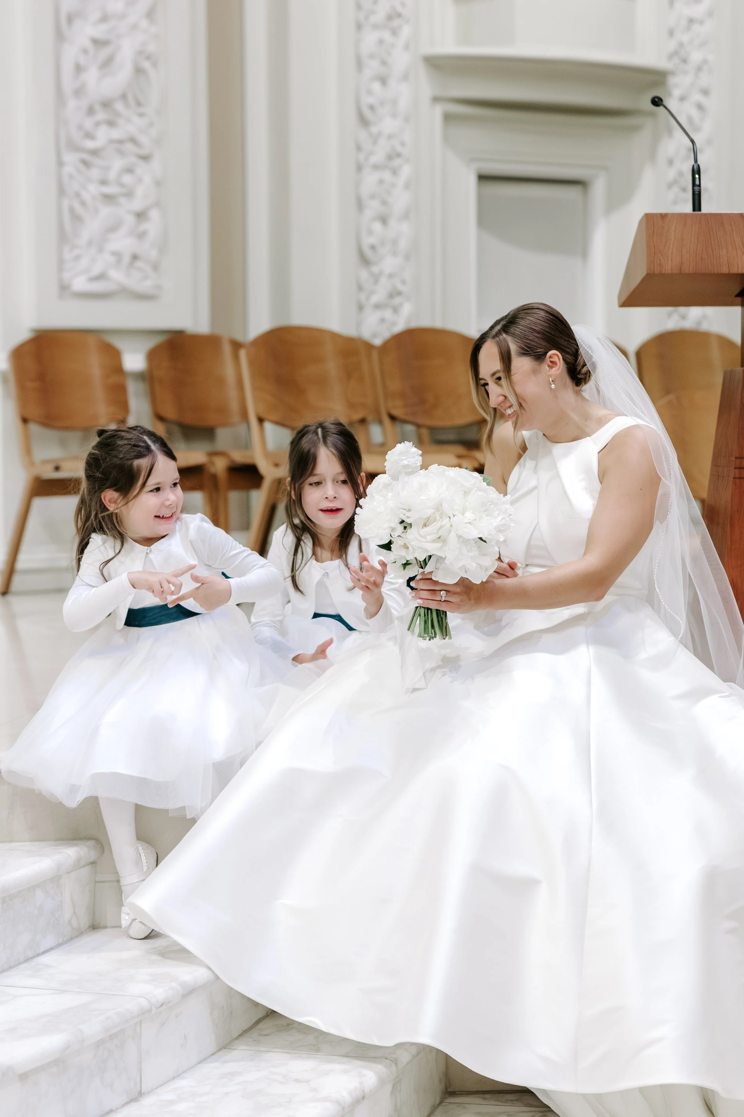 A bride in a white wedding dress sitting on white marble stairs, smiling and holding a bouquet, with two young girls in white dresses nearby, in a decorated church with white ornate walls and wooden chairs in the background.