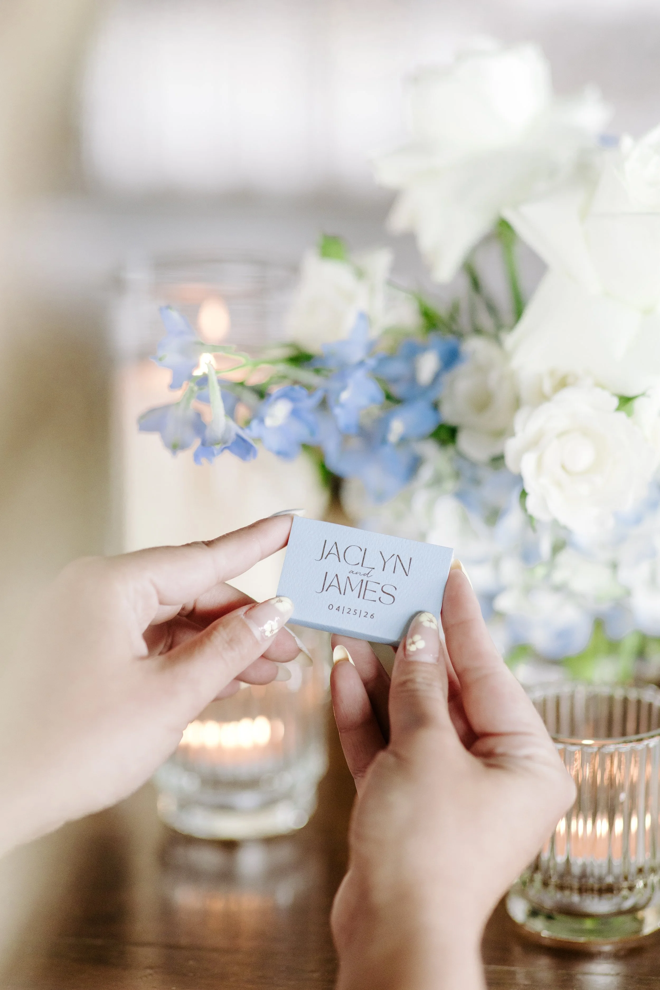 A person holding a white card with the names 'JACLYN and JAMES' and the date '04/25/26' written on it, in front of a bouquet of white and blue flowers in a glass vase, with a candle in the background.