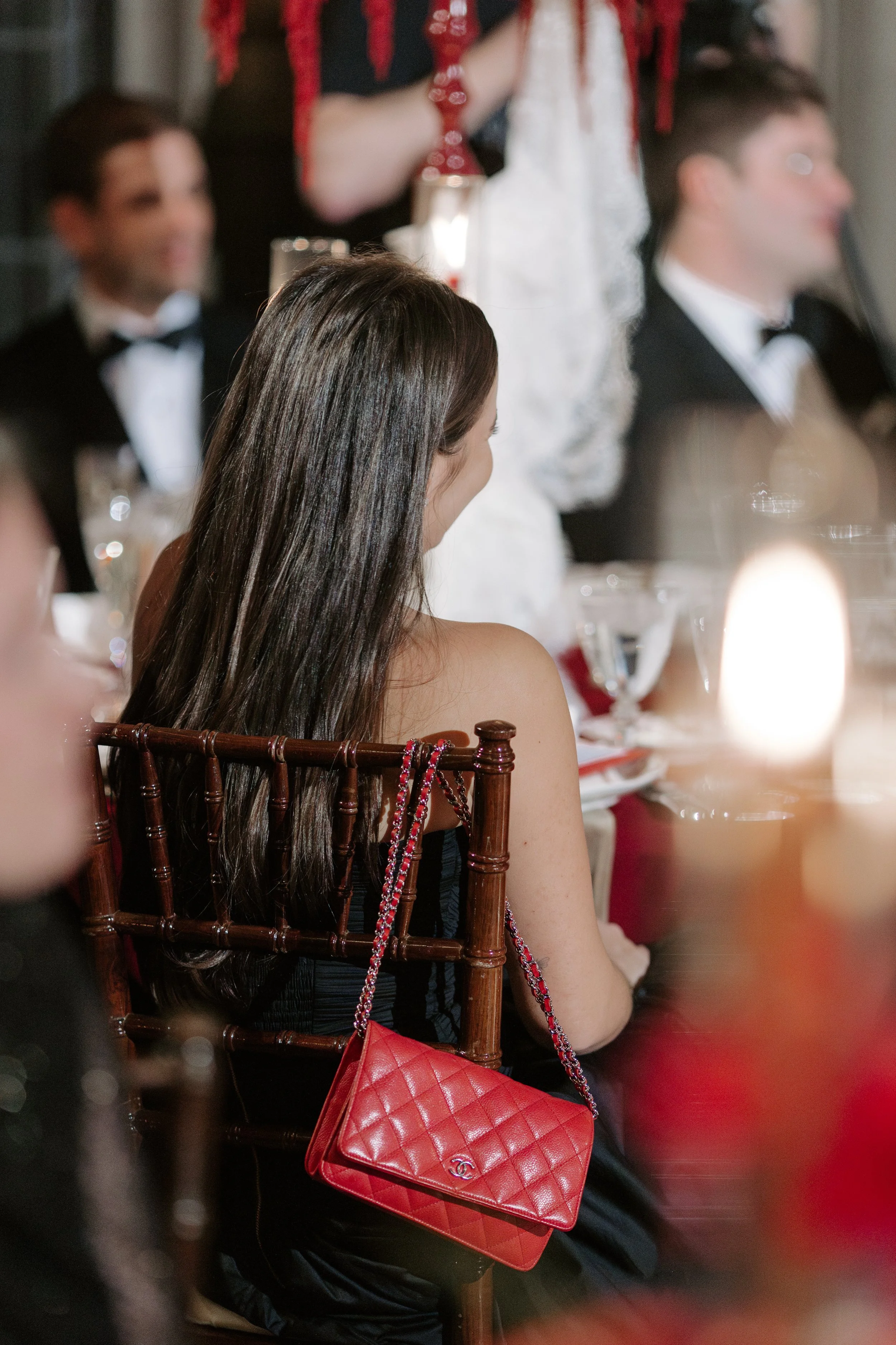 A woman with long dark hair sitting at a table during a formal event, wearing a black strapless dress and a red quilted Chanel purse with a chain strap on the back of her chair. There are blurred men in tuxedos in the background, indicating a formal 
