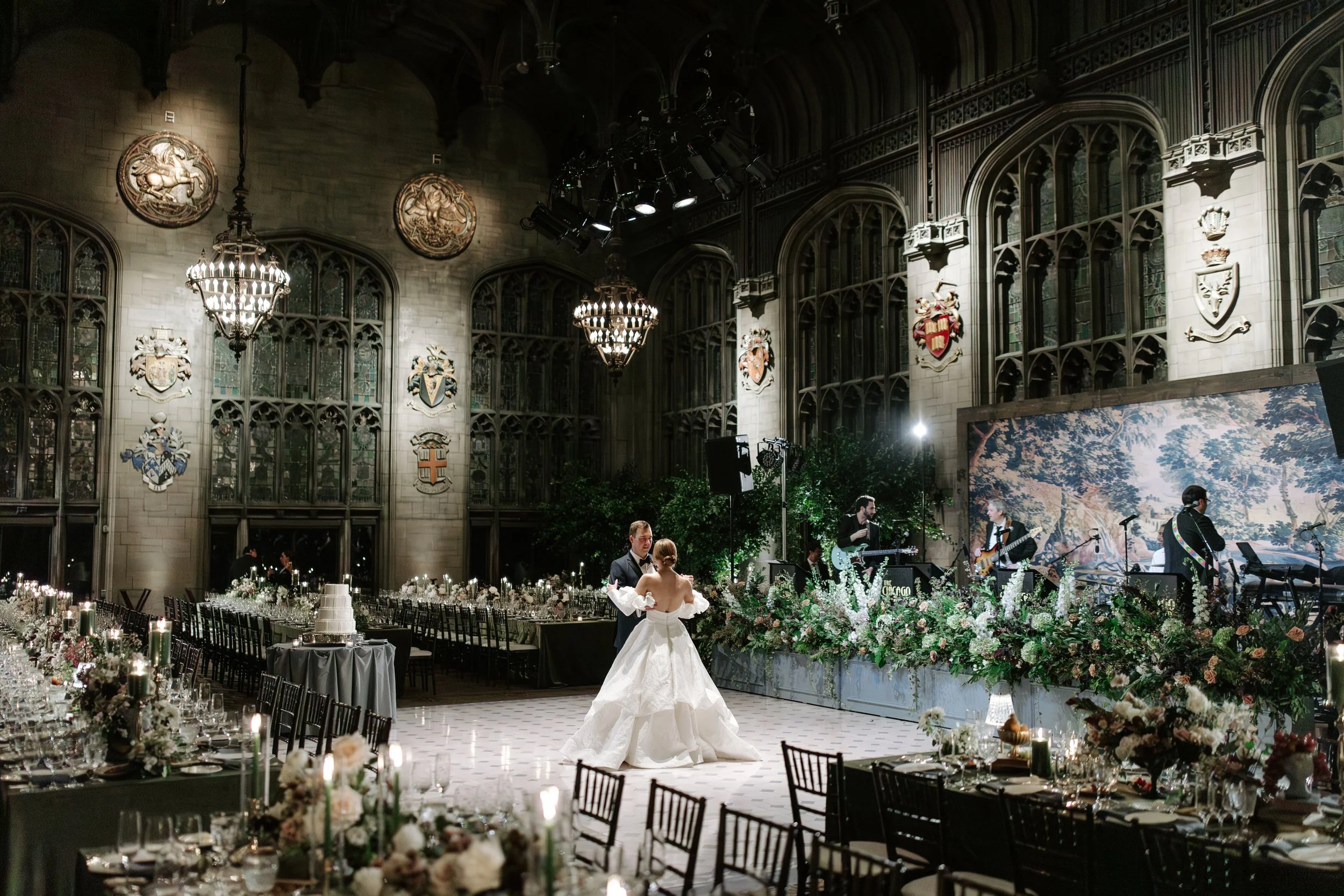 A wedding reception in a grand, historic hall with gothic architecture, chandeliers, and heraldic shields on the walls. The bride and groom are dancing, with a live band performing on stage. The tables are elegantly decorated with flowers and candles
