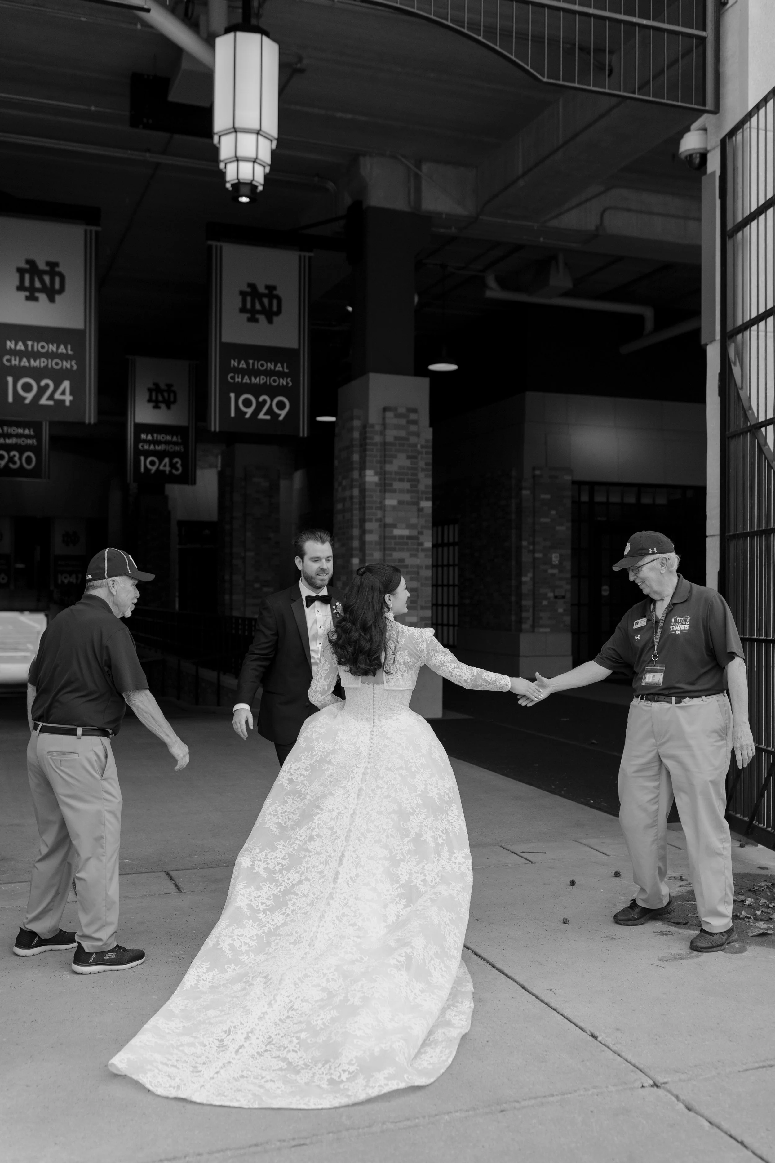 A bride in a lace wedding dress and a groom in a tuxedo holding hands with two men in casual attire, outside a stadium with championship banners hanging from the ceiling.