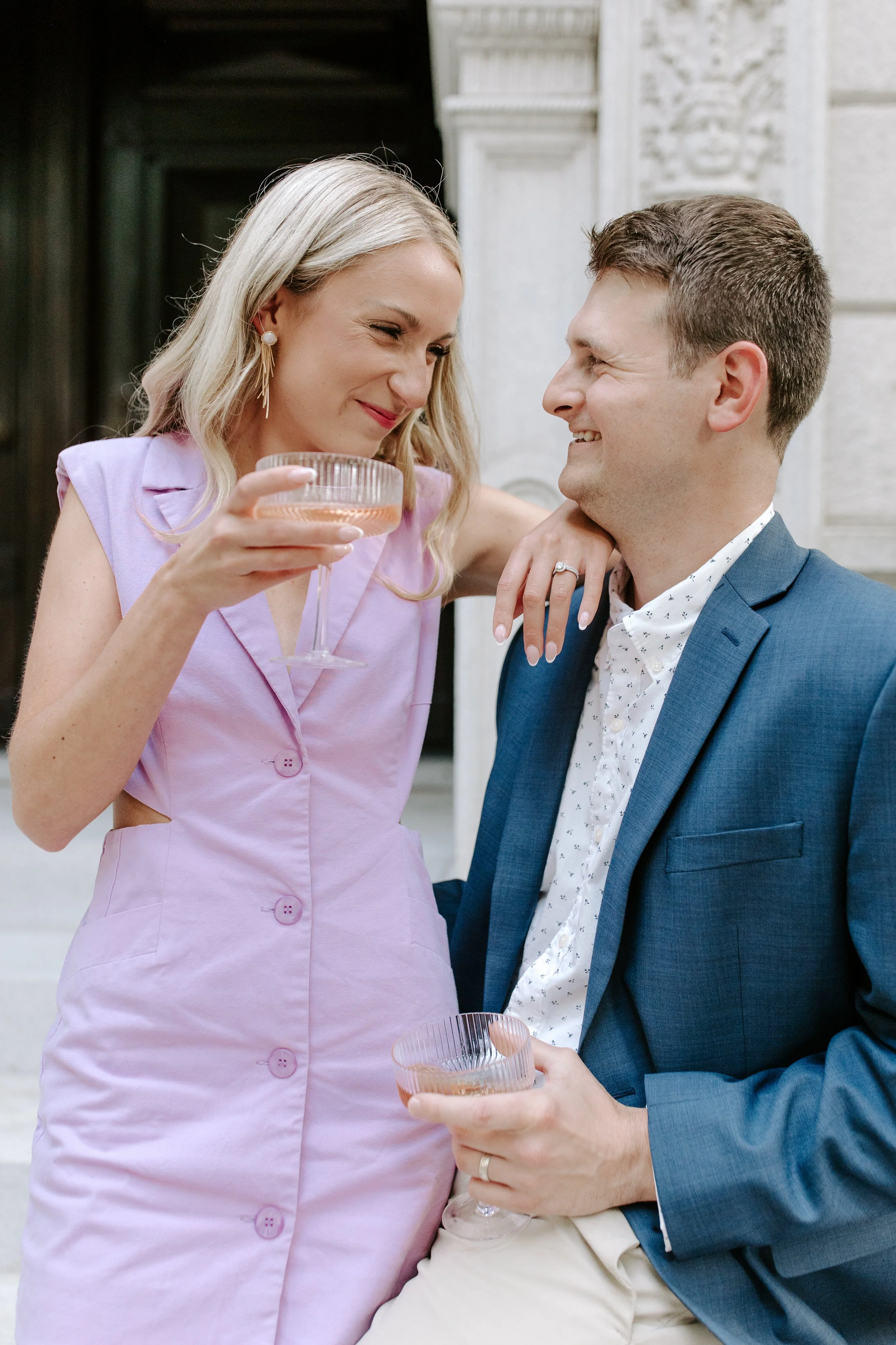 A woman in a pink dress holding a glass of pink drink, smiling at a man in a blue blazer holding a glass of pink drink, both smiling warmly, outside a building with white stone architecture.
