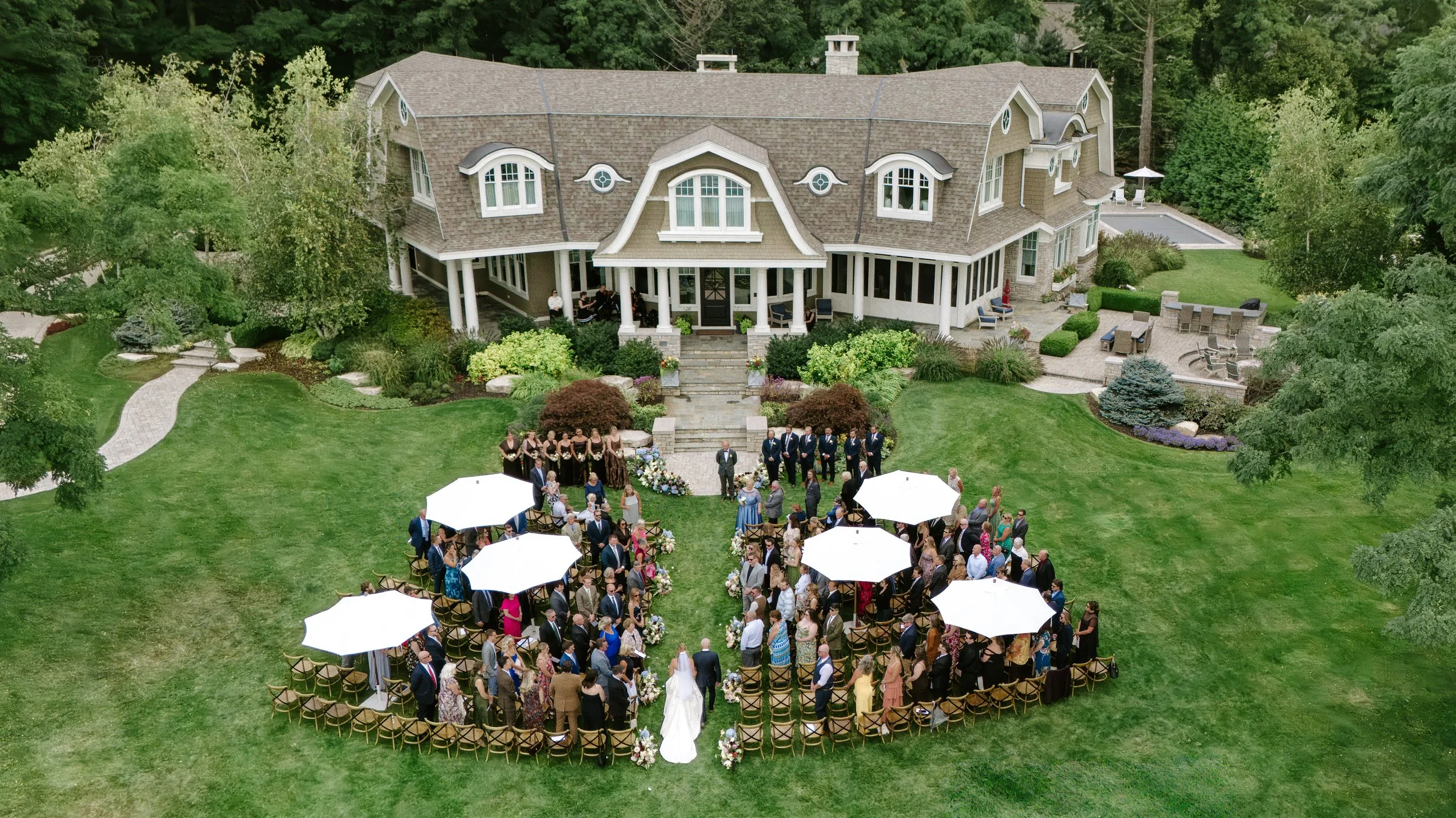An aerial view of a large house with a wedding ceremony taking place in the front yard. There are many guests seated around white umbrellas and chairs, with a bridal couple at the front near flower arrangements. The house is surrounded by greenery an
