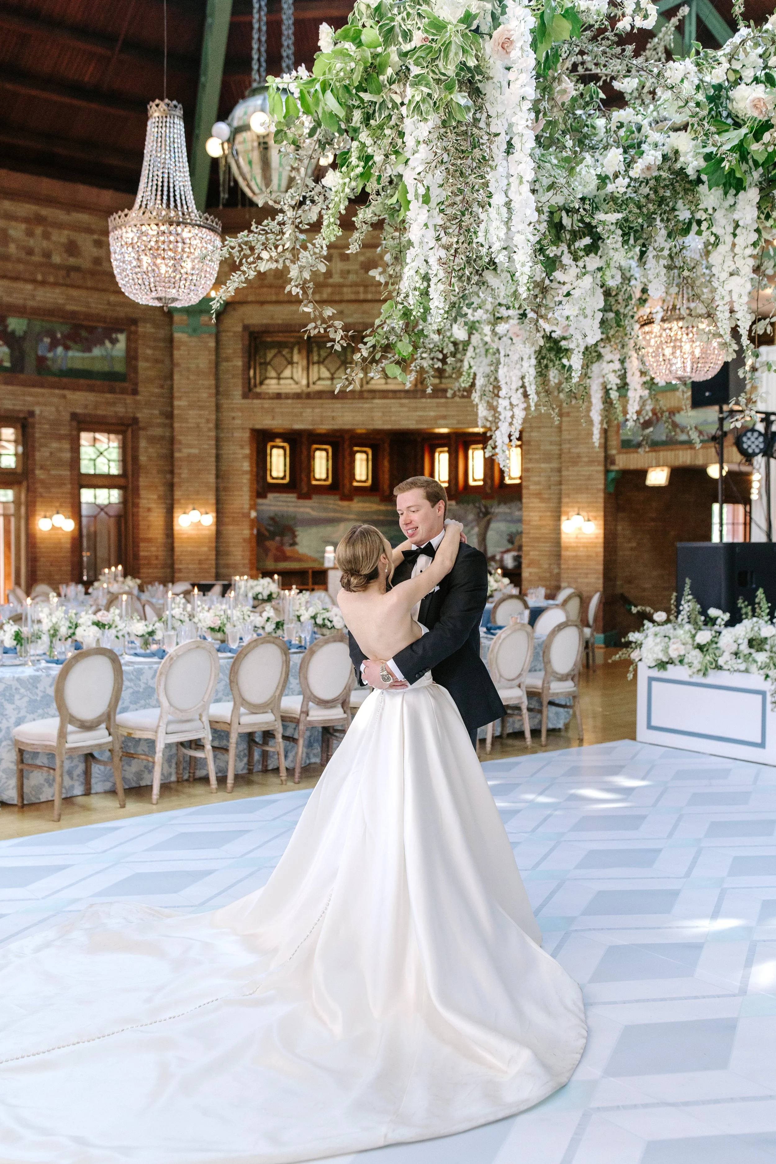 A bride and groom are dancing in a decorated wedding reception hall with white flowers hanging from the ceiling, chandeliers, and a large reception table.