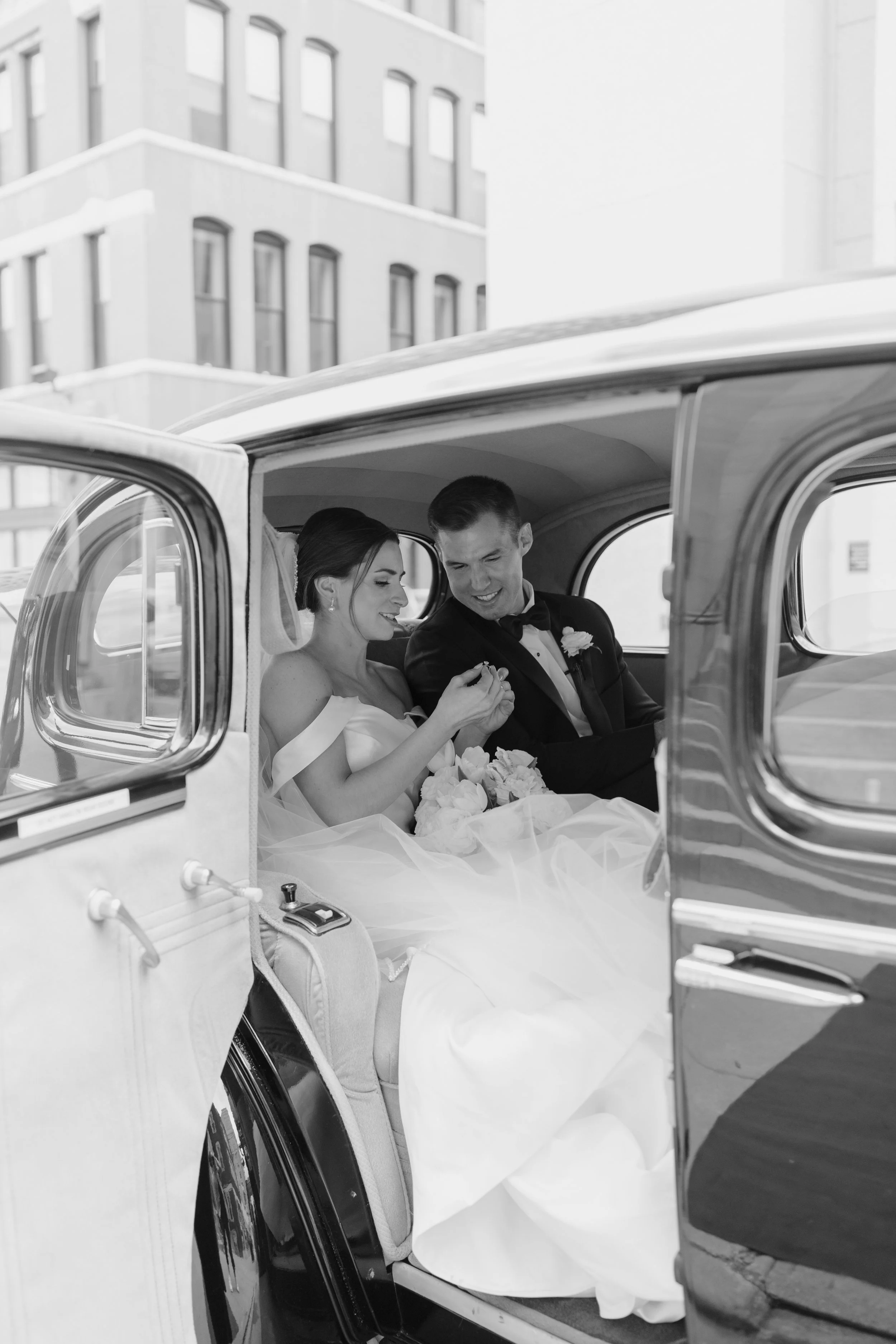 A bride and groom sitting in the back of a vintage car, smiling and looking at something. The bride is holding a bouquet of flowers and wearing a wedding dress. The groom is dressed in a tuxedo. The car's interior is visible, with the bride's veil an