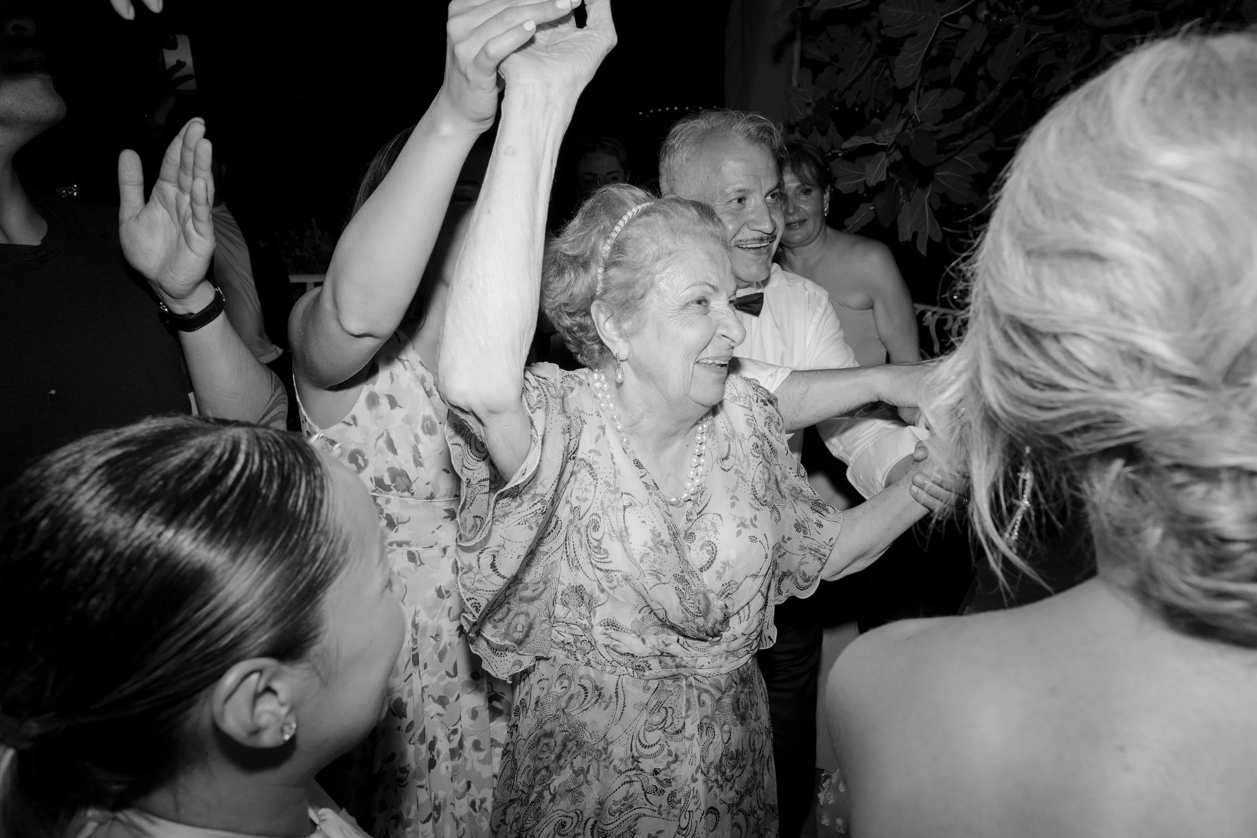 Black and white photo of an elderly woman surrounded by people at a celebratory event, holding hands and smiling.