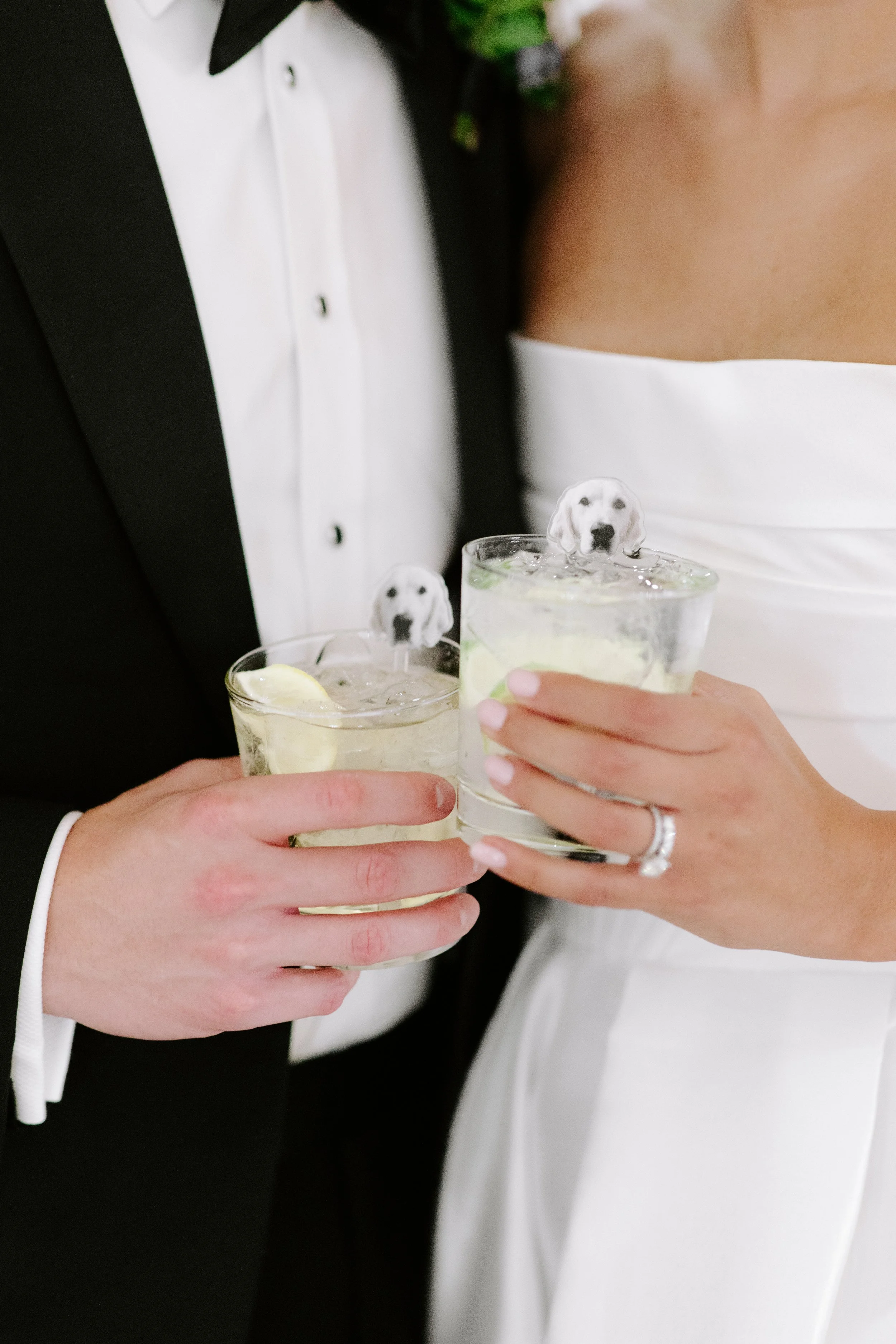 A bride and groom holding drinks with plastic puppy stir sticks in a wedding setting, the bride in a white strapless gown and the groom in a black tuxedo.