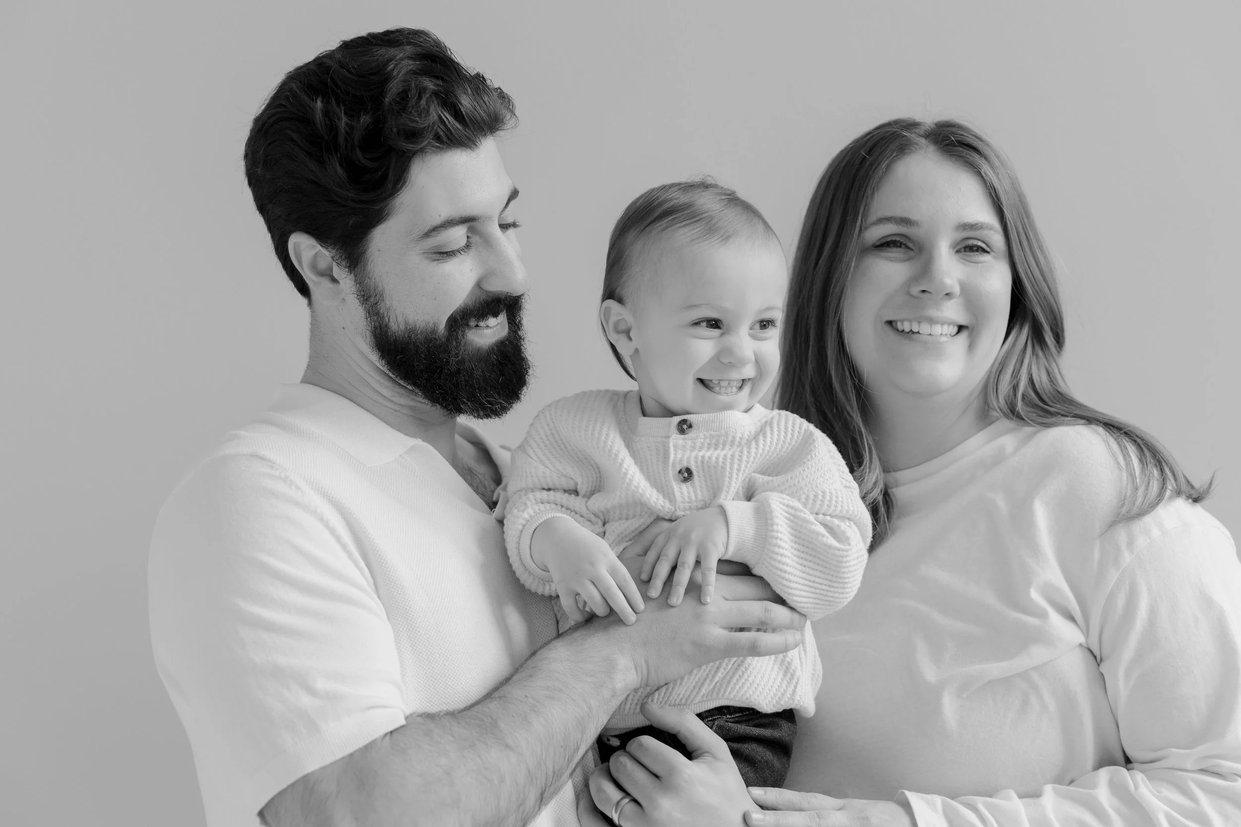 Black and white photo of a happy family of three, a man, a woman, and a young child, smiling and looking at each other with the man holding the child.