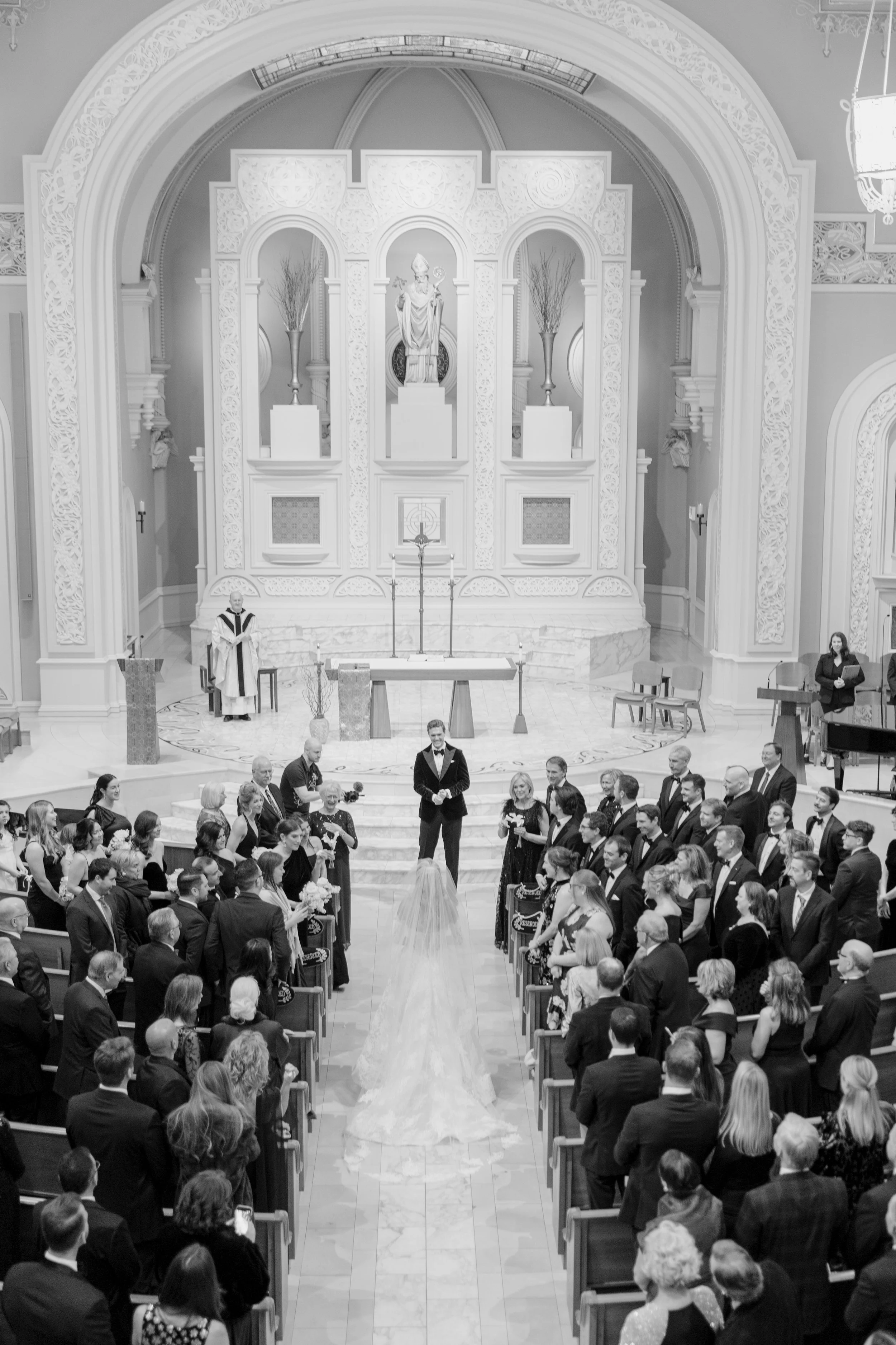 A wedding ceremony inside a church, with a bride and groom standing on the aisle, surrounded by guests seated in pews. The priest is standing to the left, and the bridal gown has a long train. The church features ornate decorations and religious stat