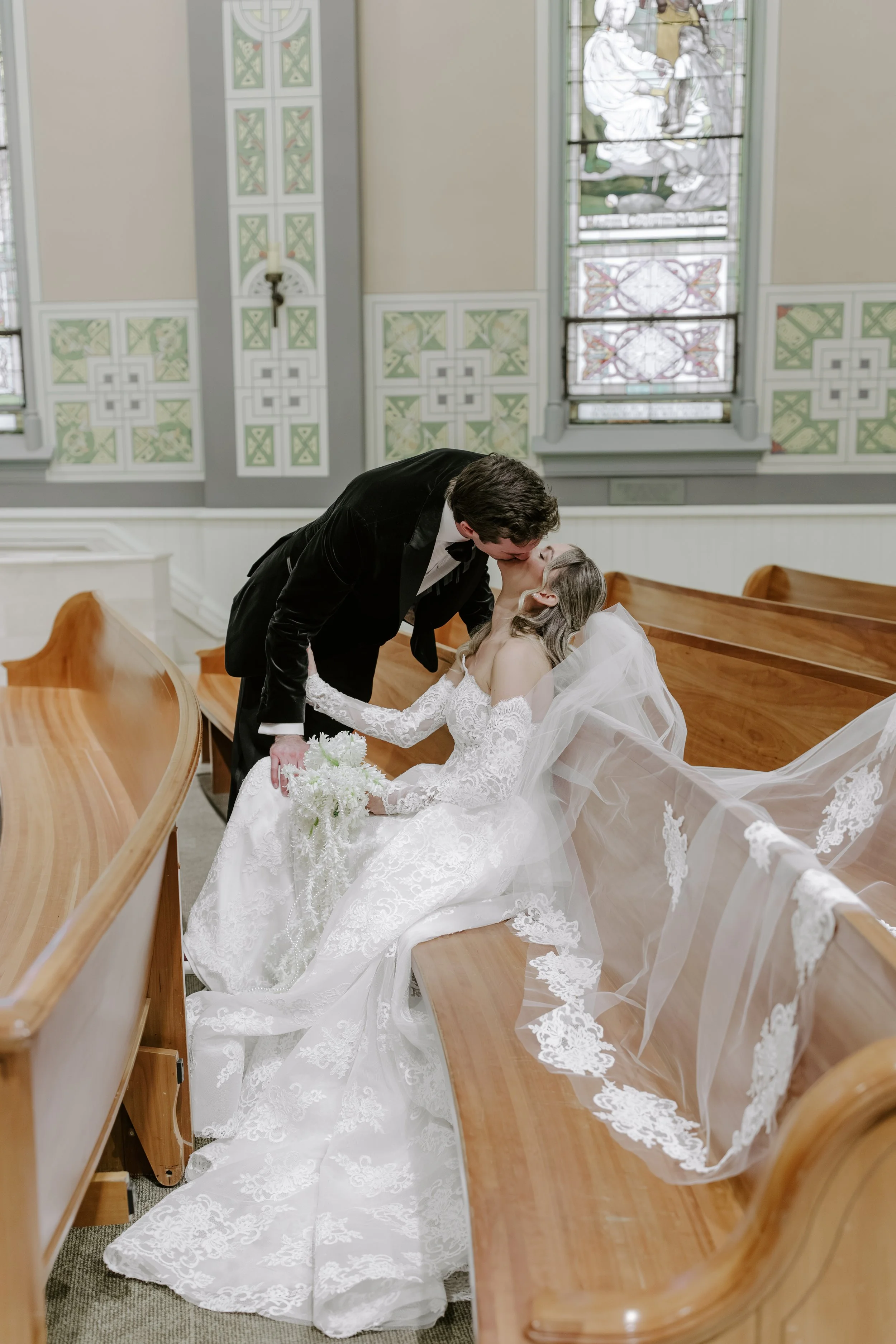 A newlywed couple sharing a kiss inside a church. The bride is seated on a pew, holding a bouquet, and wearing a white lace wedding dress and veil. The groom is standing, leaning down to kiss her, dressed in a black tuxedo with a bow tie. The church 
