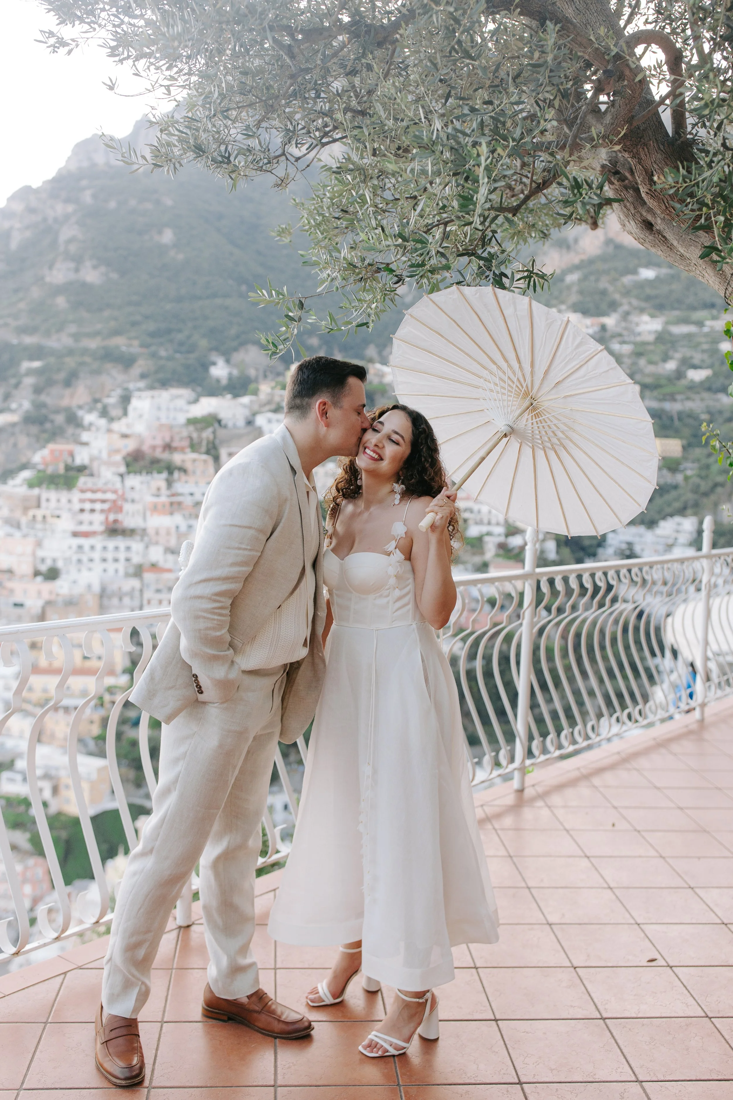 A man and a woman stand together on a balcony overlooking a hillside town. The man is wearing a light-colored suit and brown shoes, and is kissing the woman on the cheek. The woman, dressed in a white dress and white heels, is smiling and holding a w