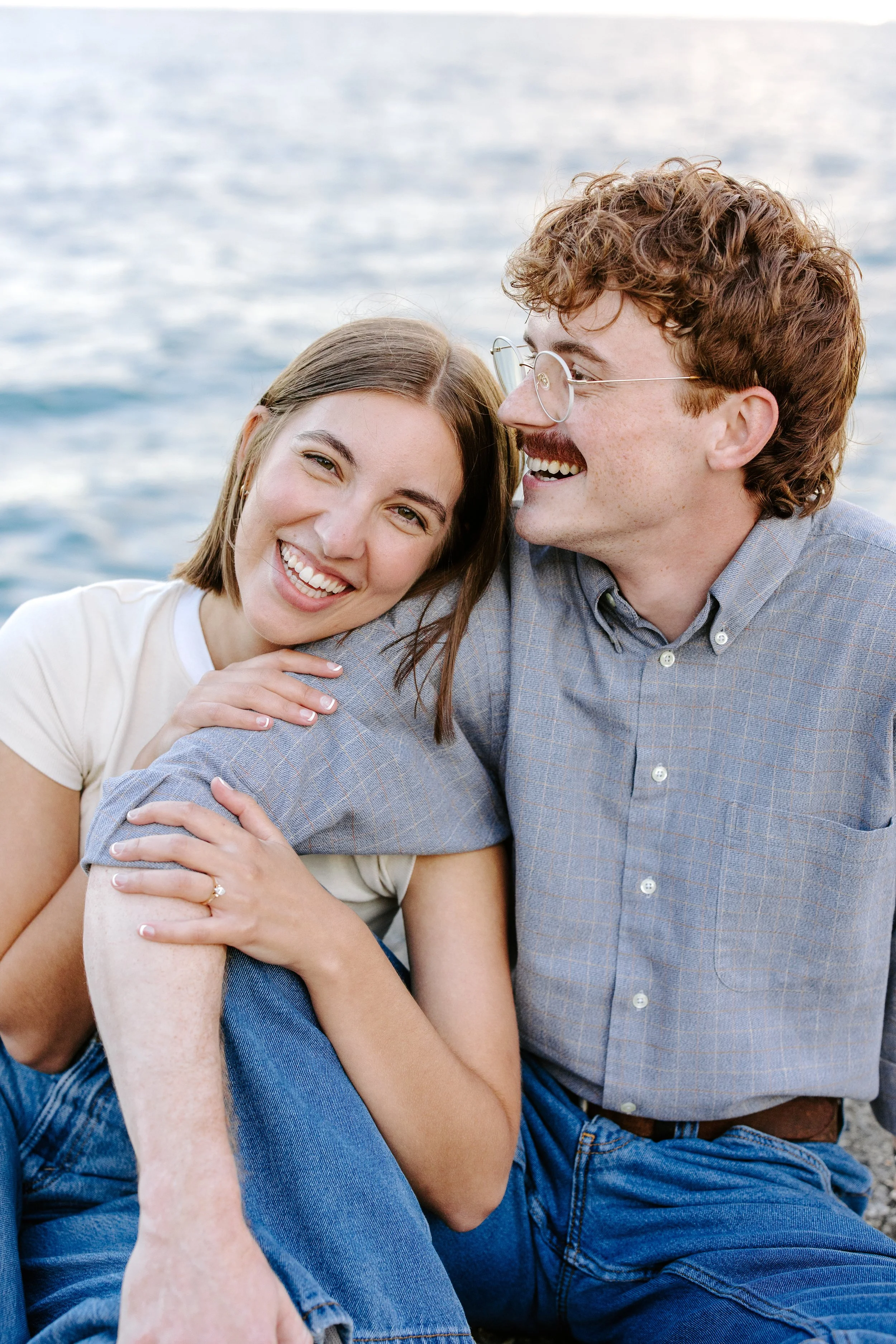 A young woman and a young man sitting closely by the water, smiling and sharing a joyful moment together.