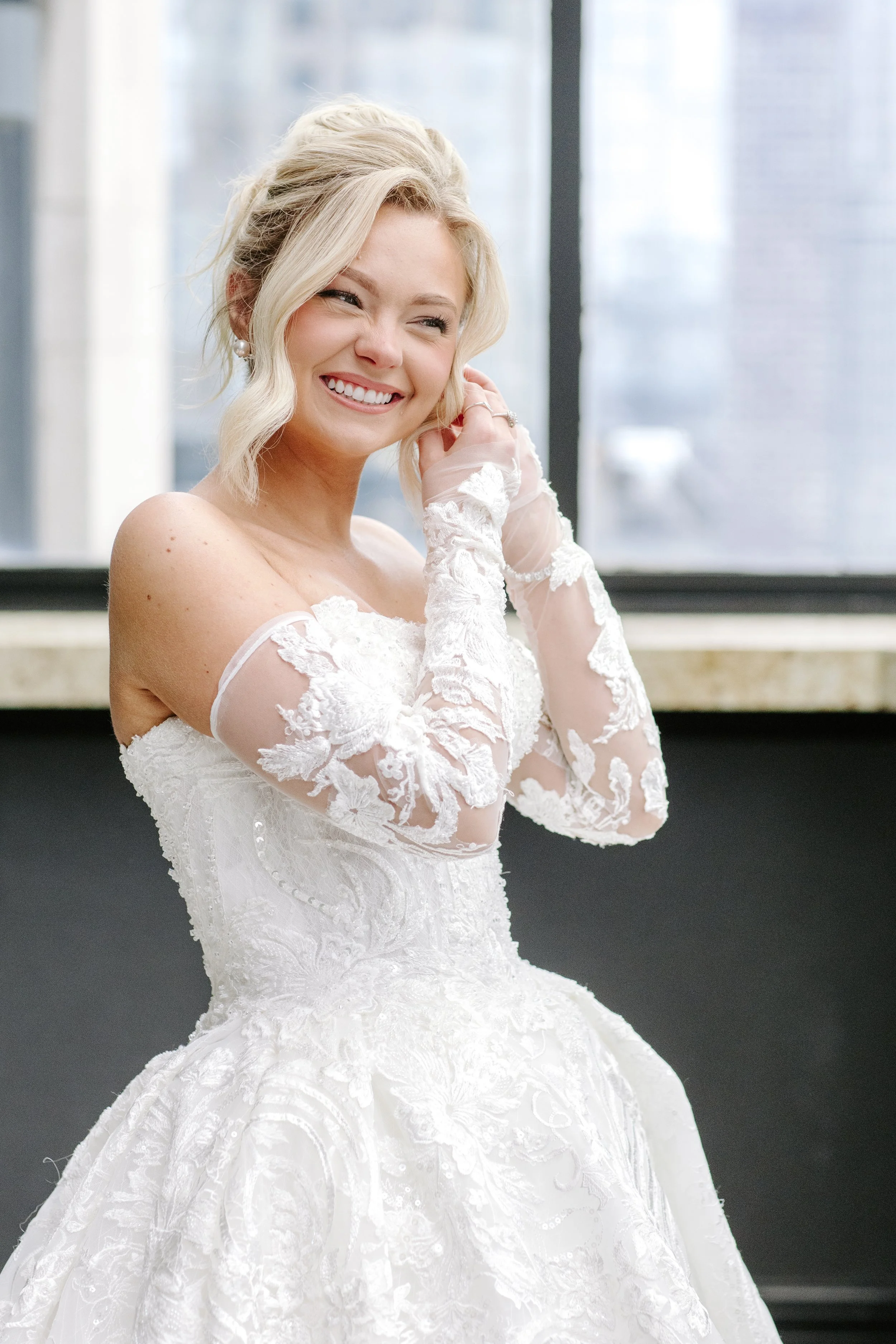A woman in a white wedding dress smiling and adjusting her earring near a window.