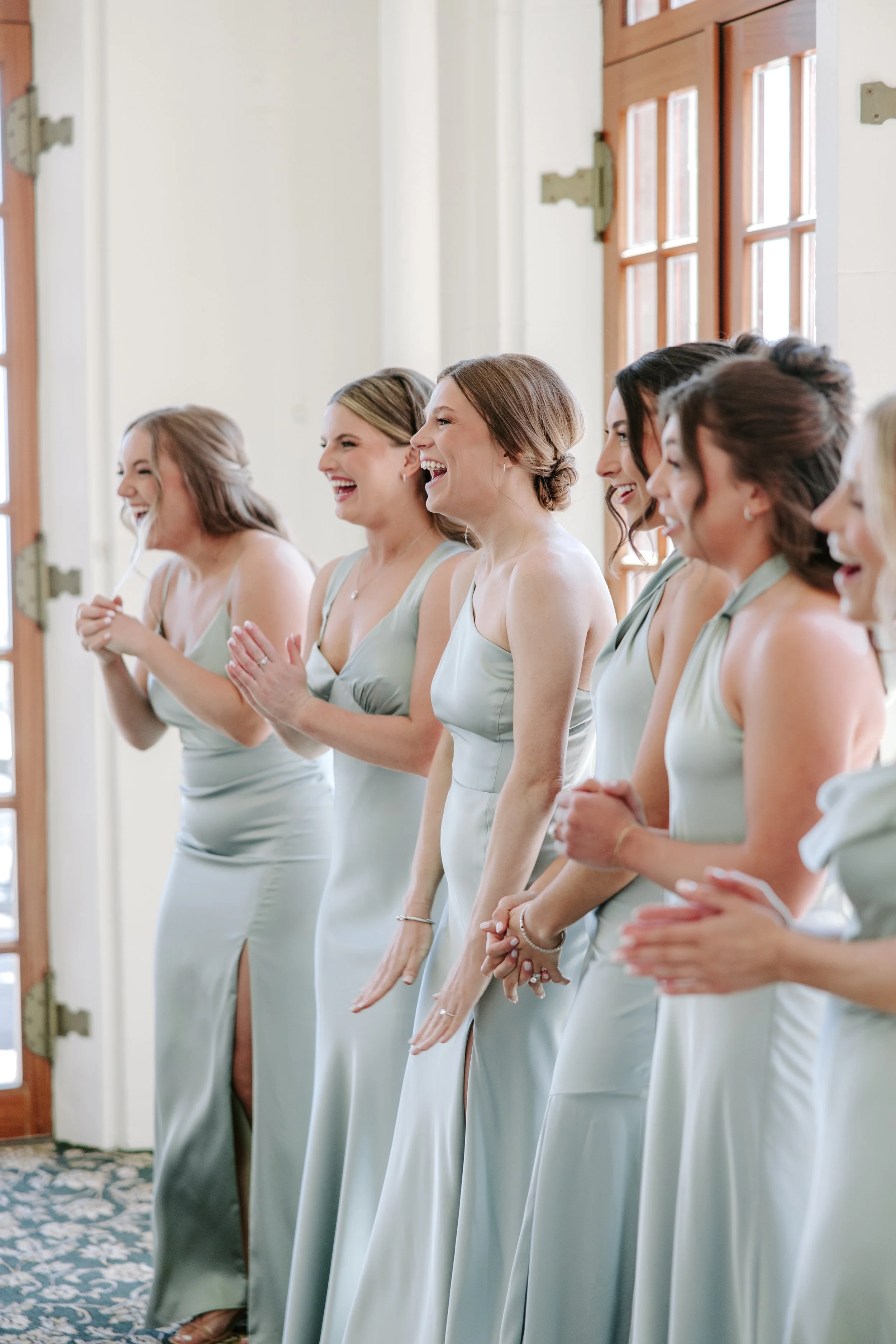 A group of women in matching pale green dresses, smiling and clapping, standing inside near windows with natural light.