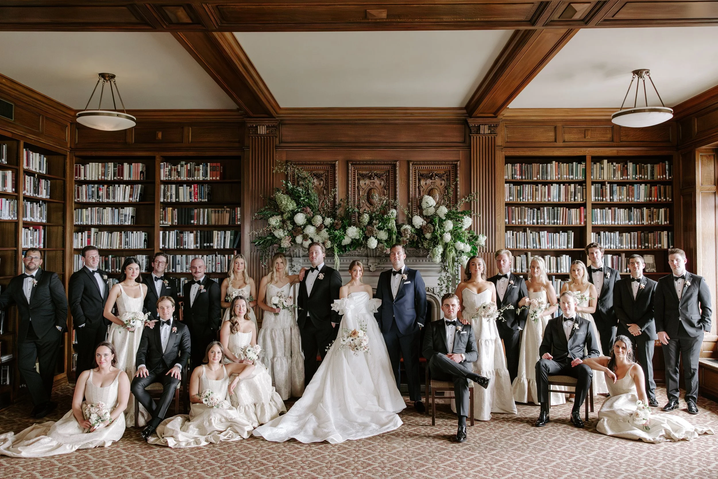A large group of wedding party members, including bridesmaids and groomsmen, posing for a formal photo in a wood-paneled library with high bookshelves and large floral arrangements.