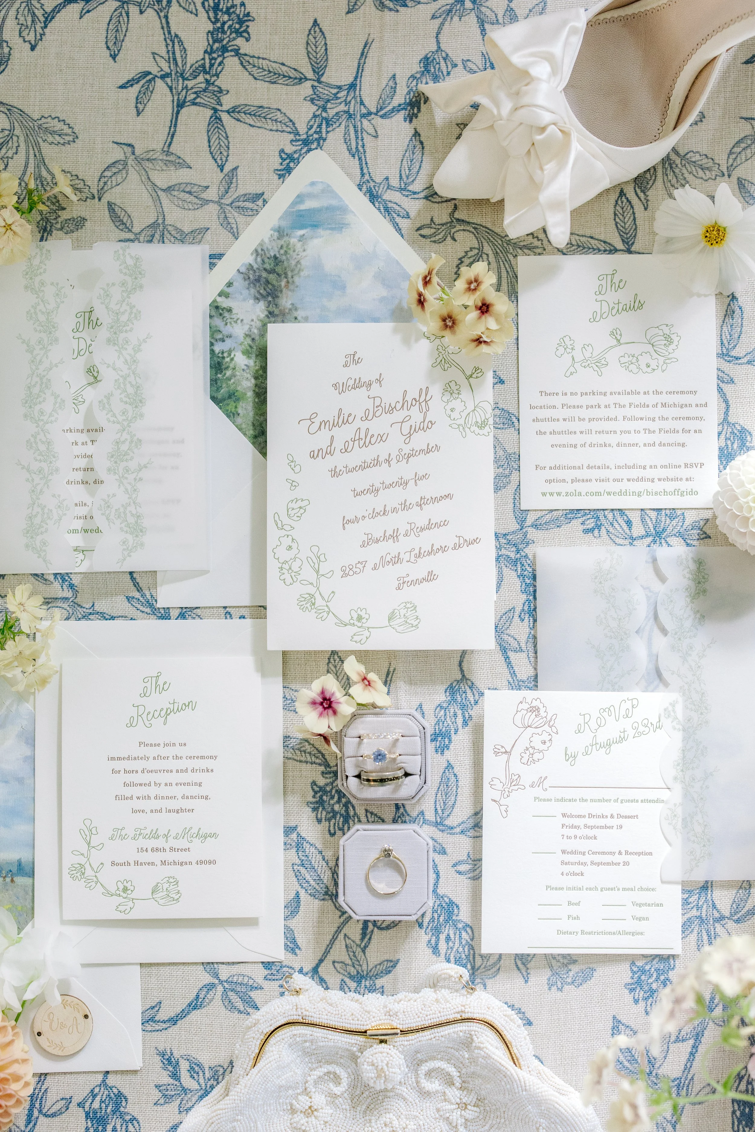 Wedding invitation cards, wedding rings, flowers, and shoes on a blue and white patterned tablecloth.