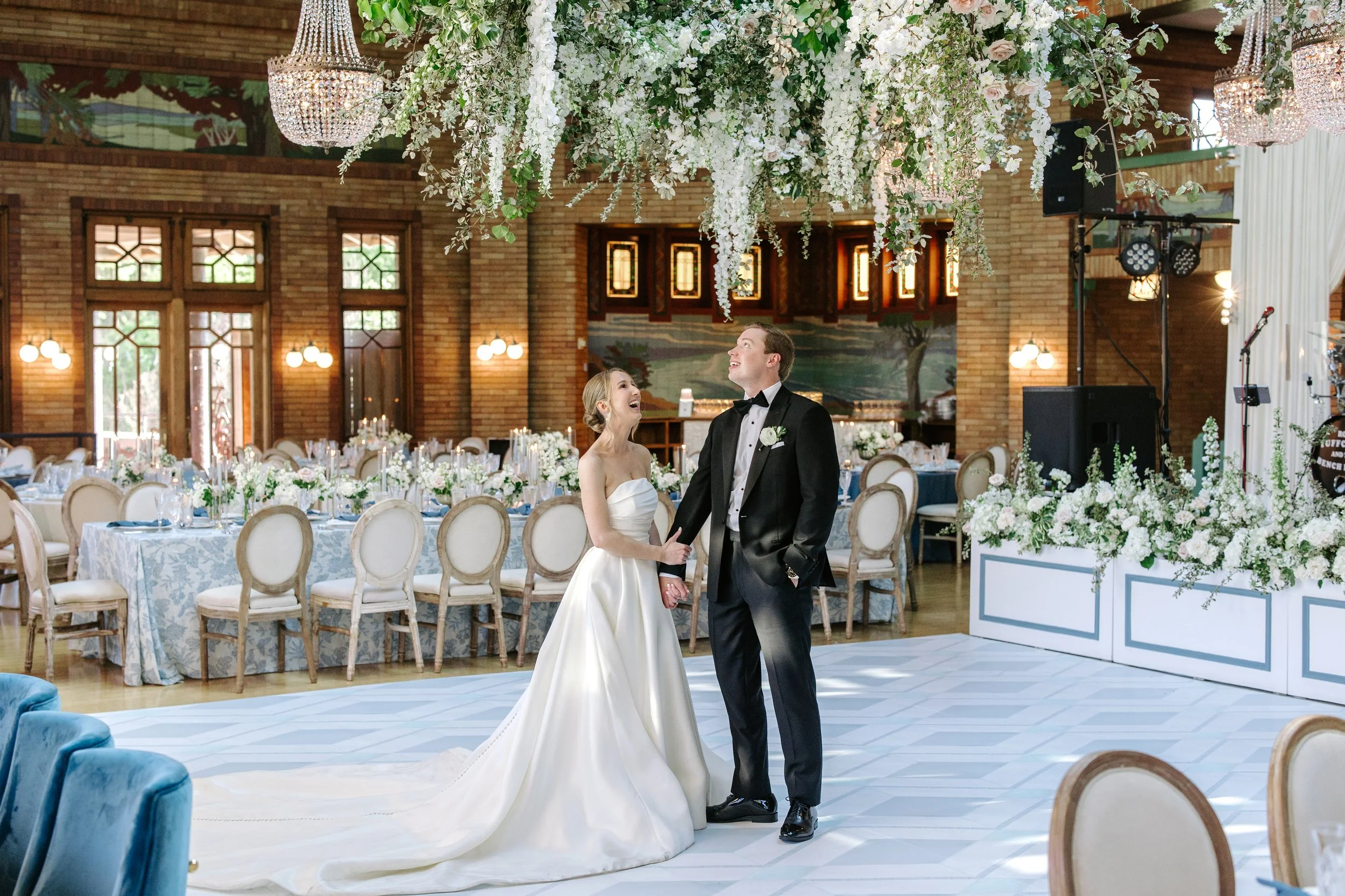 A bride and groom dance together at their wedding reception in a decorated banquet hall filled with tables, chairs, floral arrangements, and chandeliers.