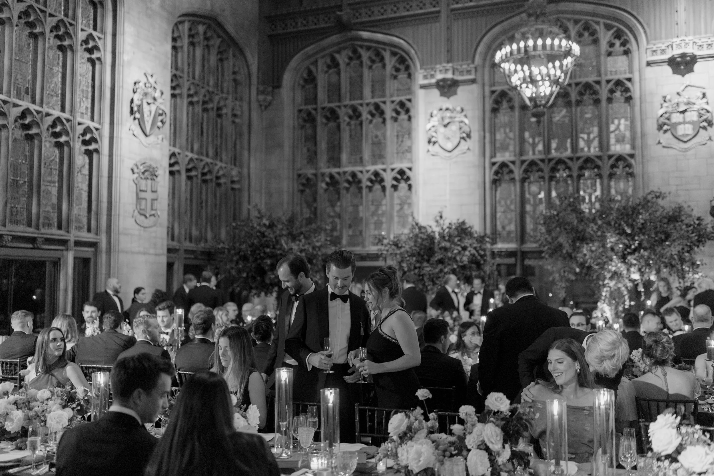 A black and white photograph of a formal event inside a grand historic hall with high vaulted ceilings, large stained glass windows, and ornate decorations on the walls. Multiple guests in tuxedos and evening gowns are engaged in conversations, and t