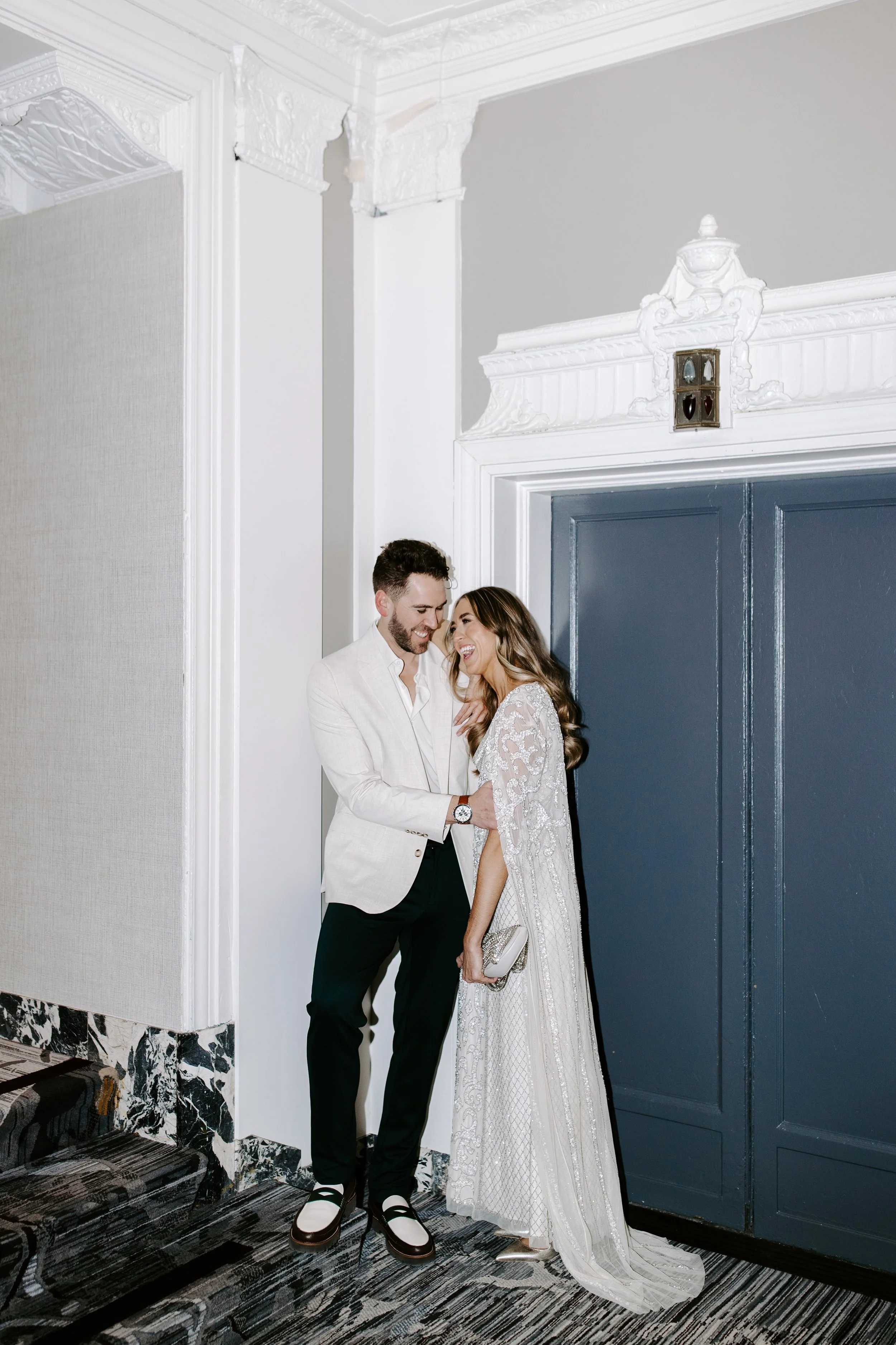 A smiling man and woman dressed formally, standing in an elegant room with ornate white crown molding, a blue door, and marble flooring, sharing a happy moment.