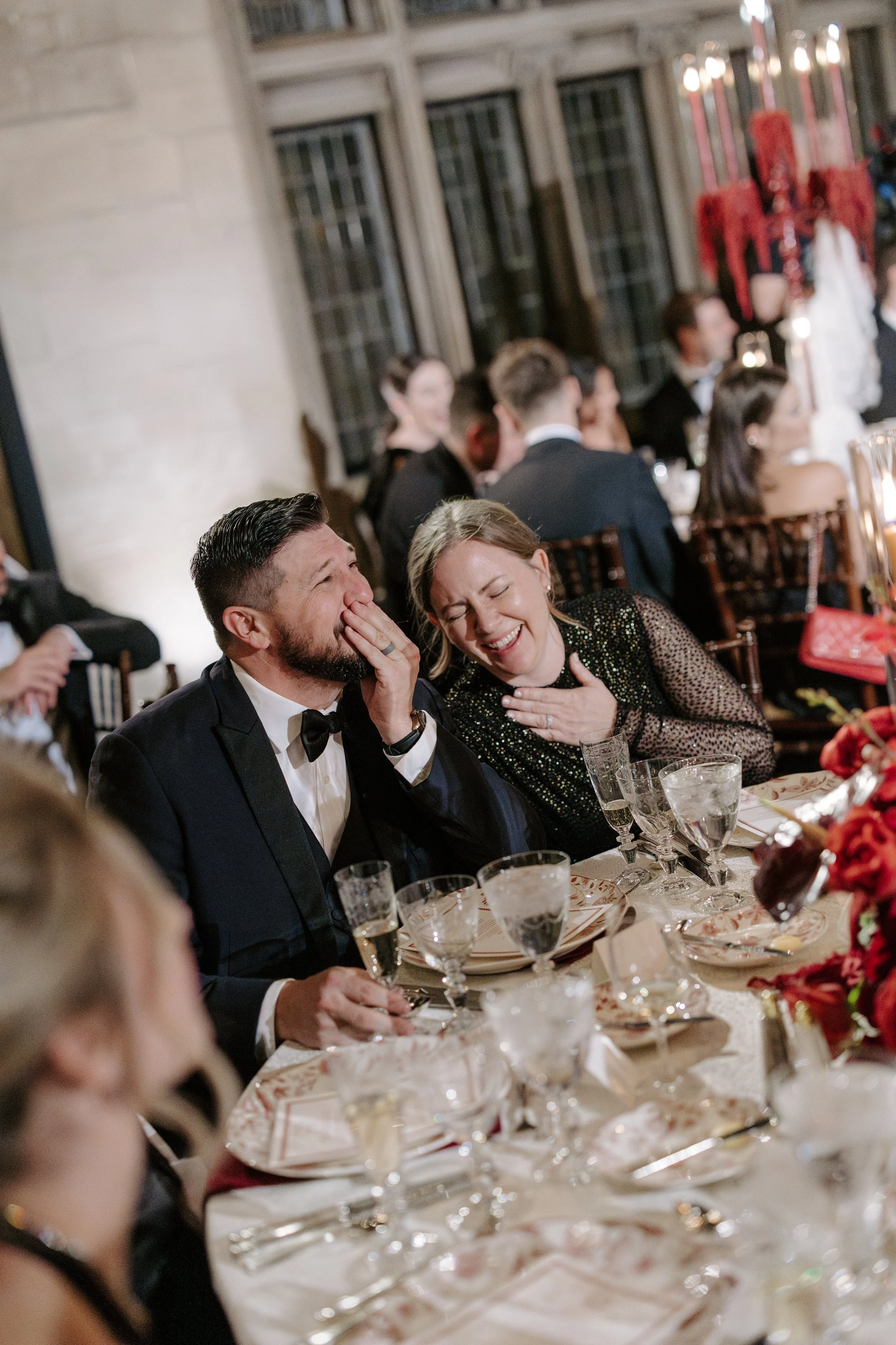 People celebrating at a formal dinner event, seated at a decorated table with glassware, flowers, and candles, dressed in formal attire, with some laughing and engaging in conversation.