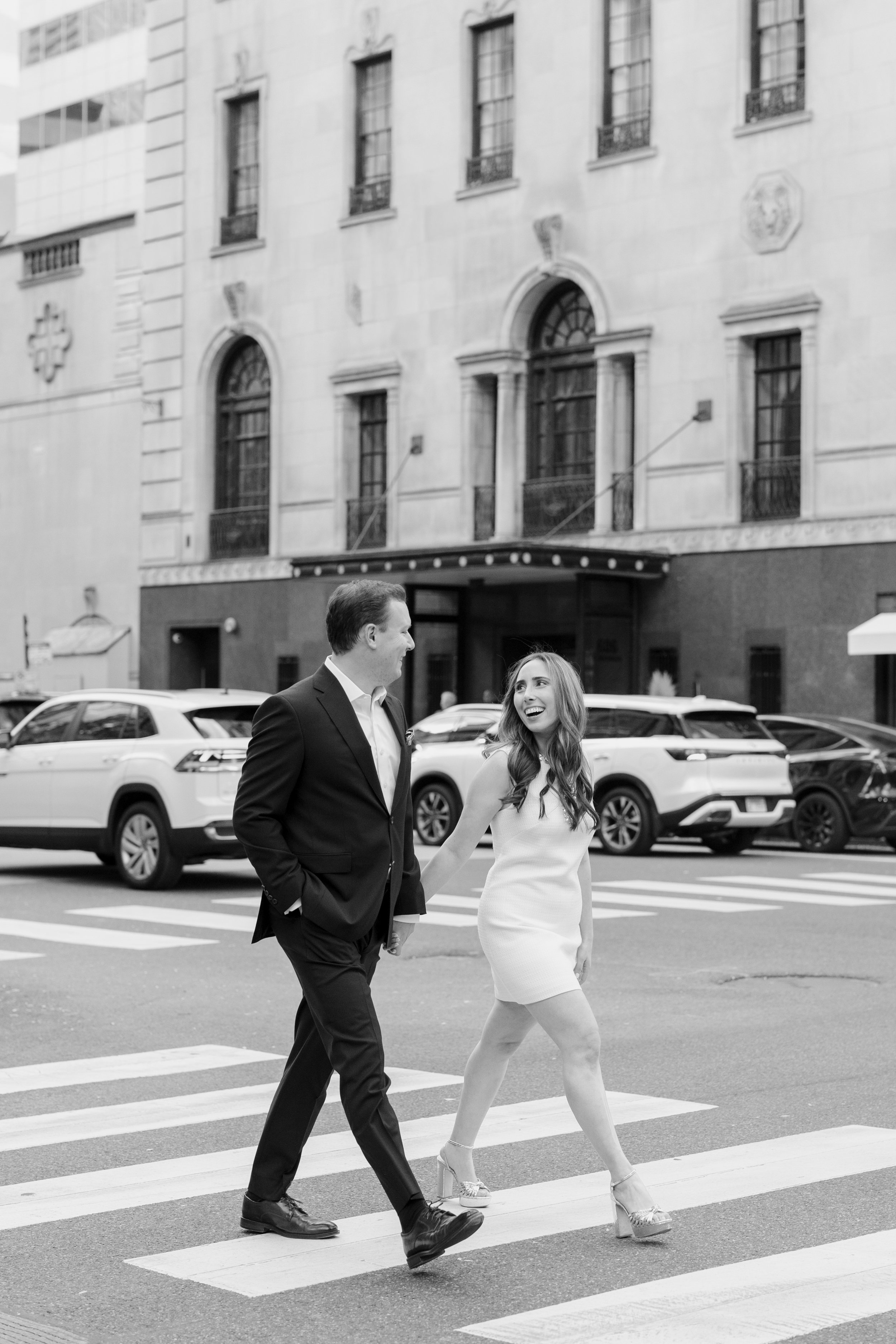 A man in a black suit and a woman in a white dress are walking hand in hand across a crosswalk in an urban area with cars parked on the street and tall buildings in the background.