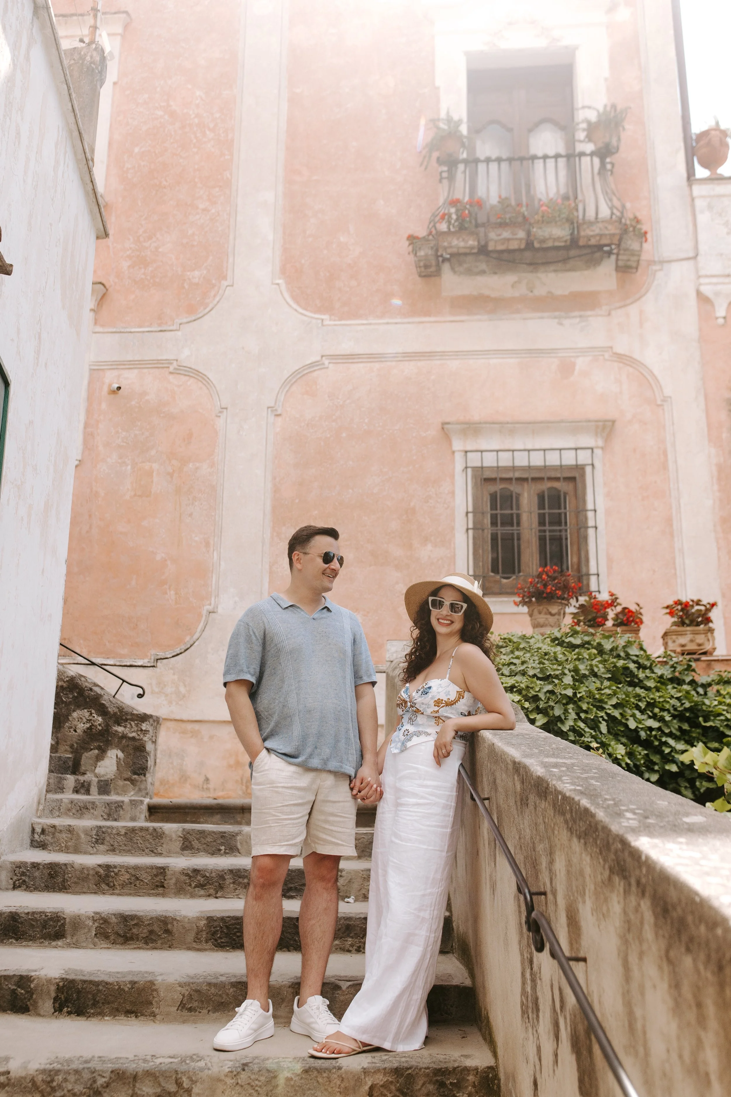 A man and woman standing on stone stairs outside a building, holding hands. The man is wearing a light blue shirt, beige shorts, and white sneakers. The woman is wearing a white strapless dress, a wide-brimmed hat, and sunglasses. They are smiling an