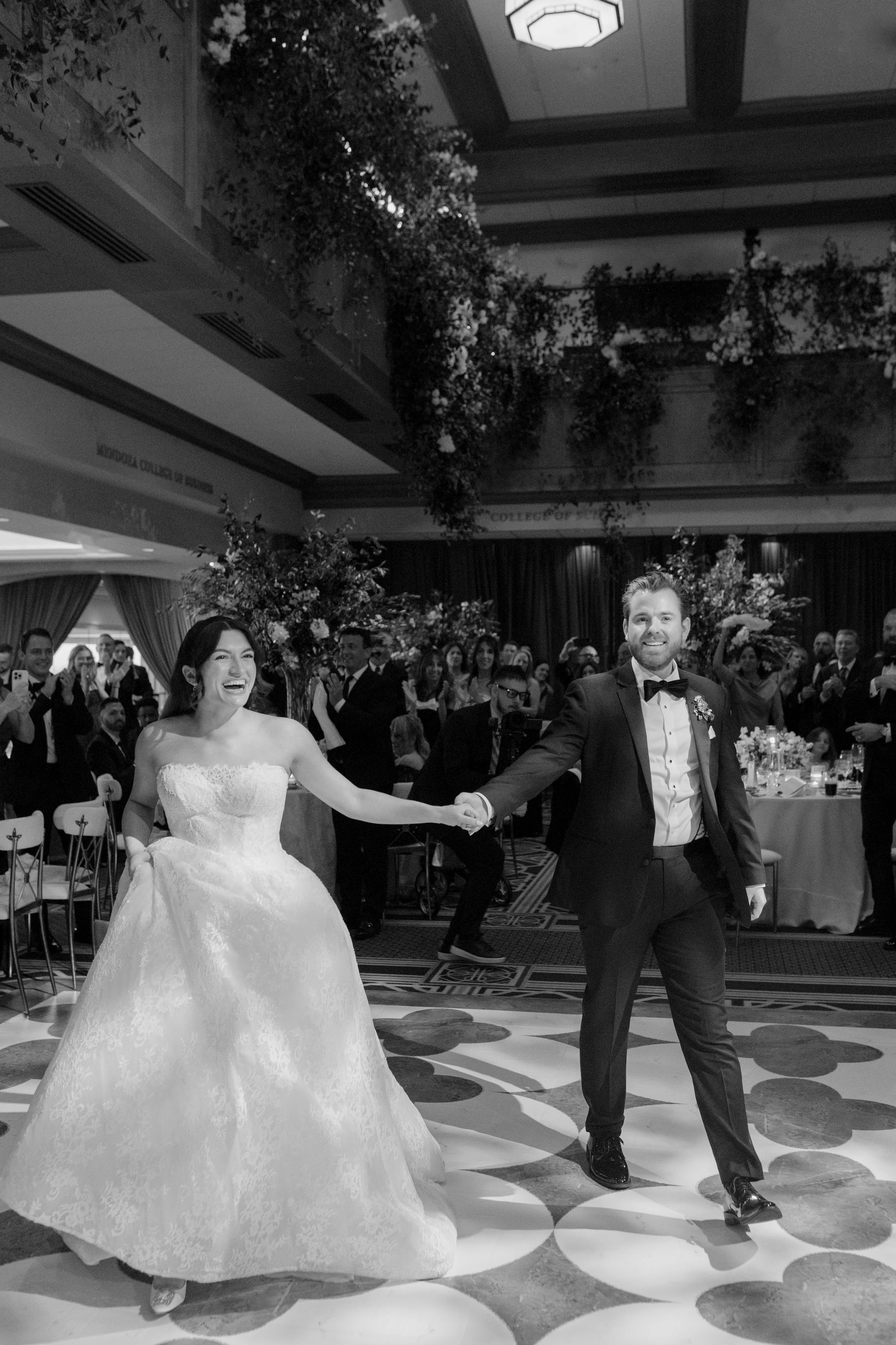 A newlywed couple dancing at their wedding reception, with guests in formal attire watching and clapping around them. The bride is in a strapless wedding gown, and the groom is in a tuxedo with a bow tie, in an elegant decorated banquet hall.
