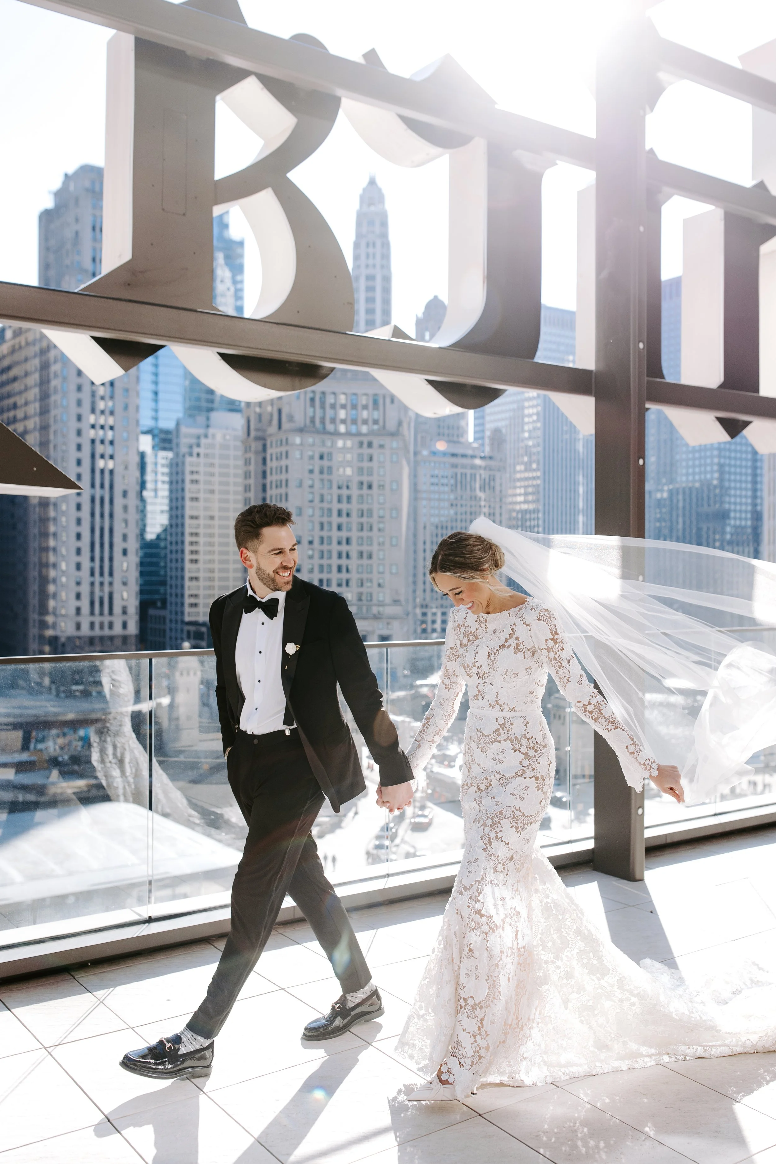 A bride and groom walking hand in hand on a rooftop terrace with a city skyline background, smiling and dressed in wedding attire.