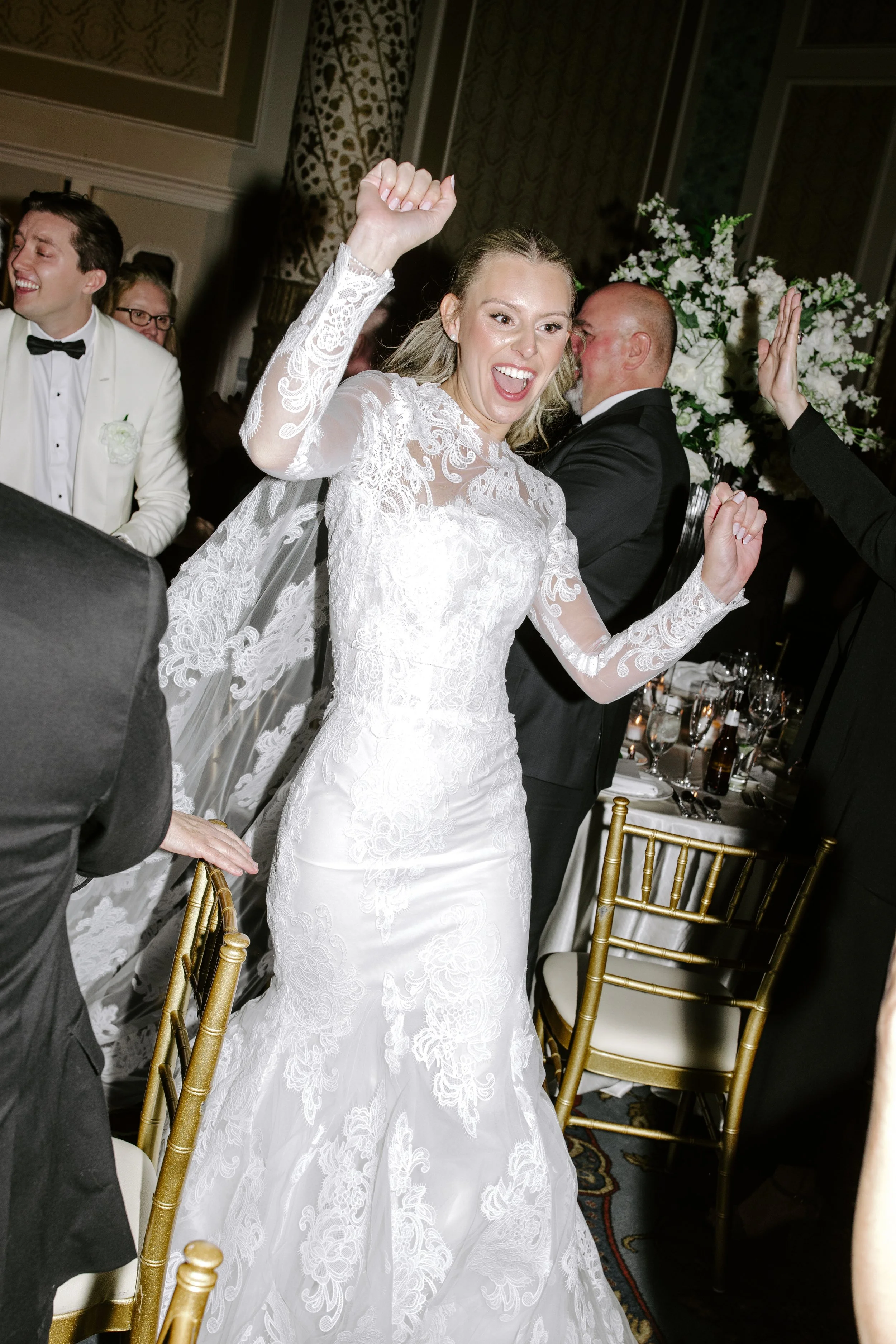 A bride in a white lace wedding dress celebrating at her wedding reception with her arms raised and friends around her.