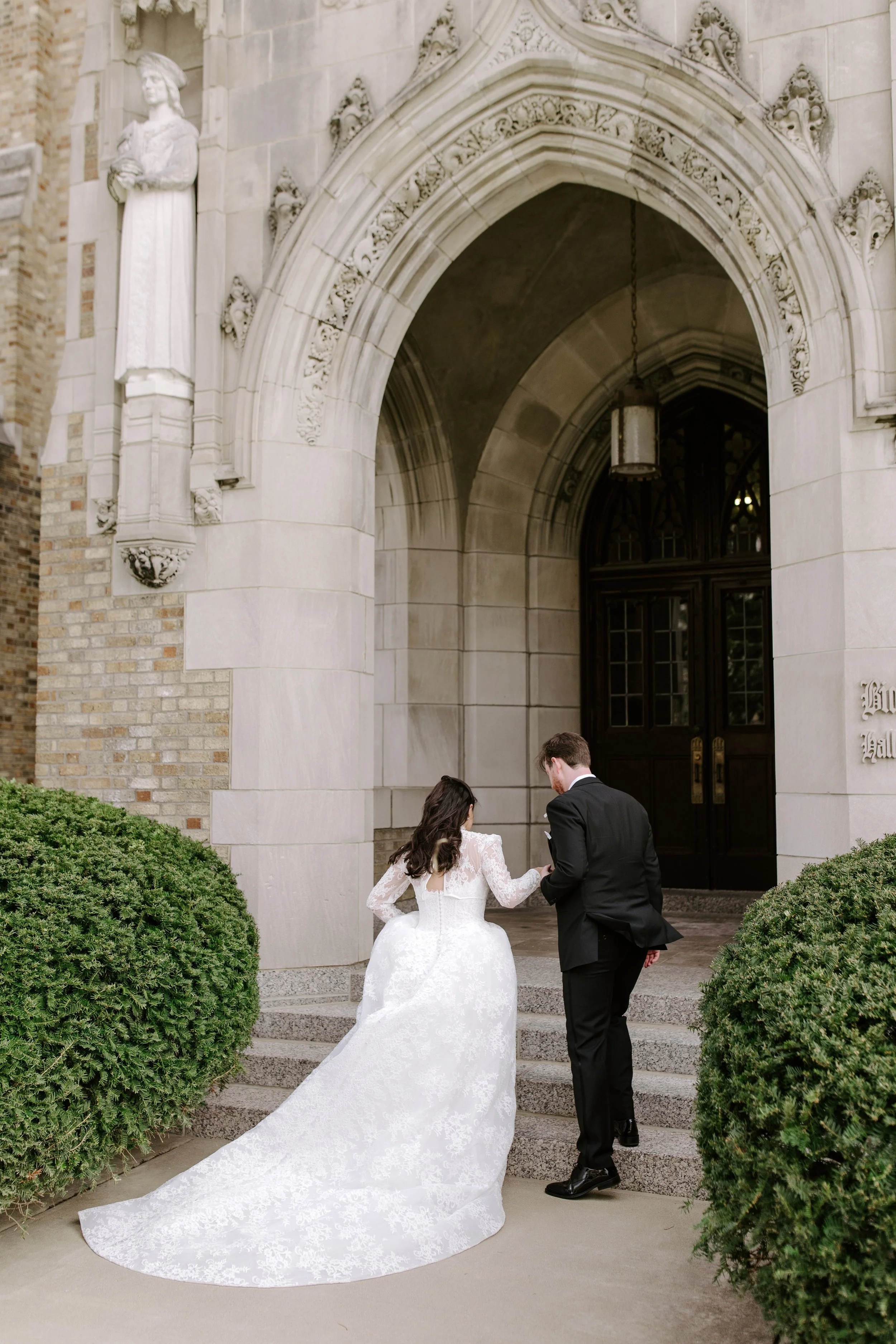 A bride and groom standing on steps outside a gothic-style church, holding hands, with the bride wearing a white wedding gown with a long train and the groom in a black suit.