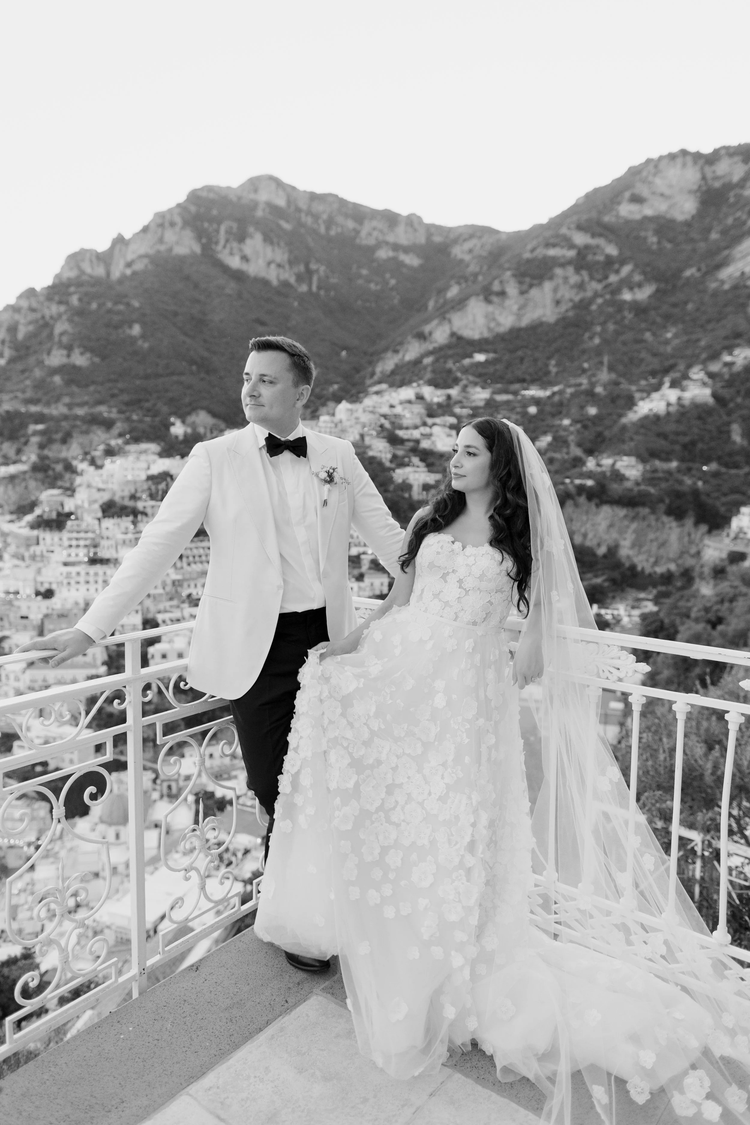 Black and white photo of a bride and groom on a balcony with a mountainous landscape in the background. The groom is in a white tuxedo with a black bow tie, looking to the left. The bride is in a lace wedding gown with a long veil, sitting, and looki