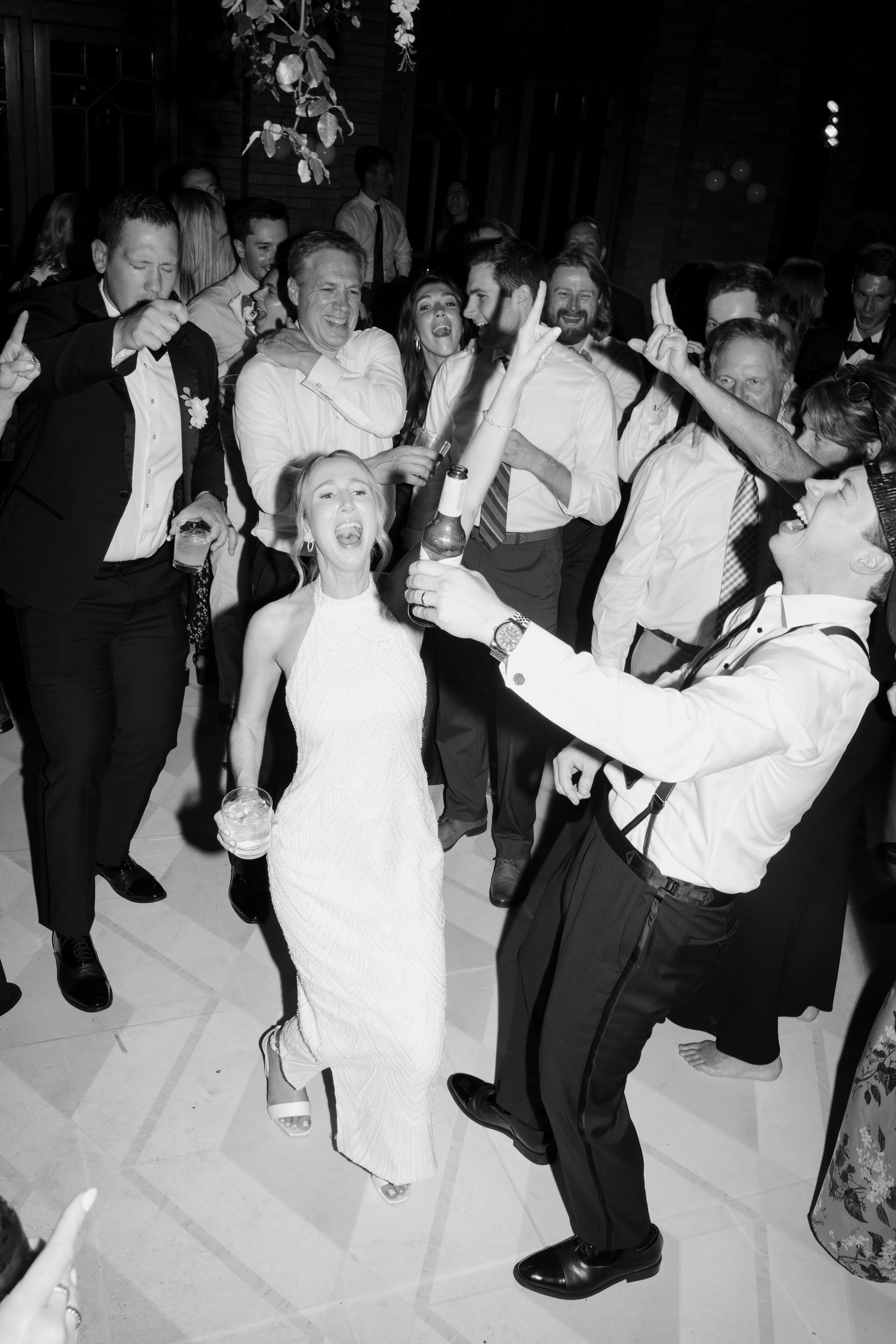 A group of people dancing and celebrating at a party, some holding drinks, with a young girl in the foreground singing, all dressed in formal attire.