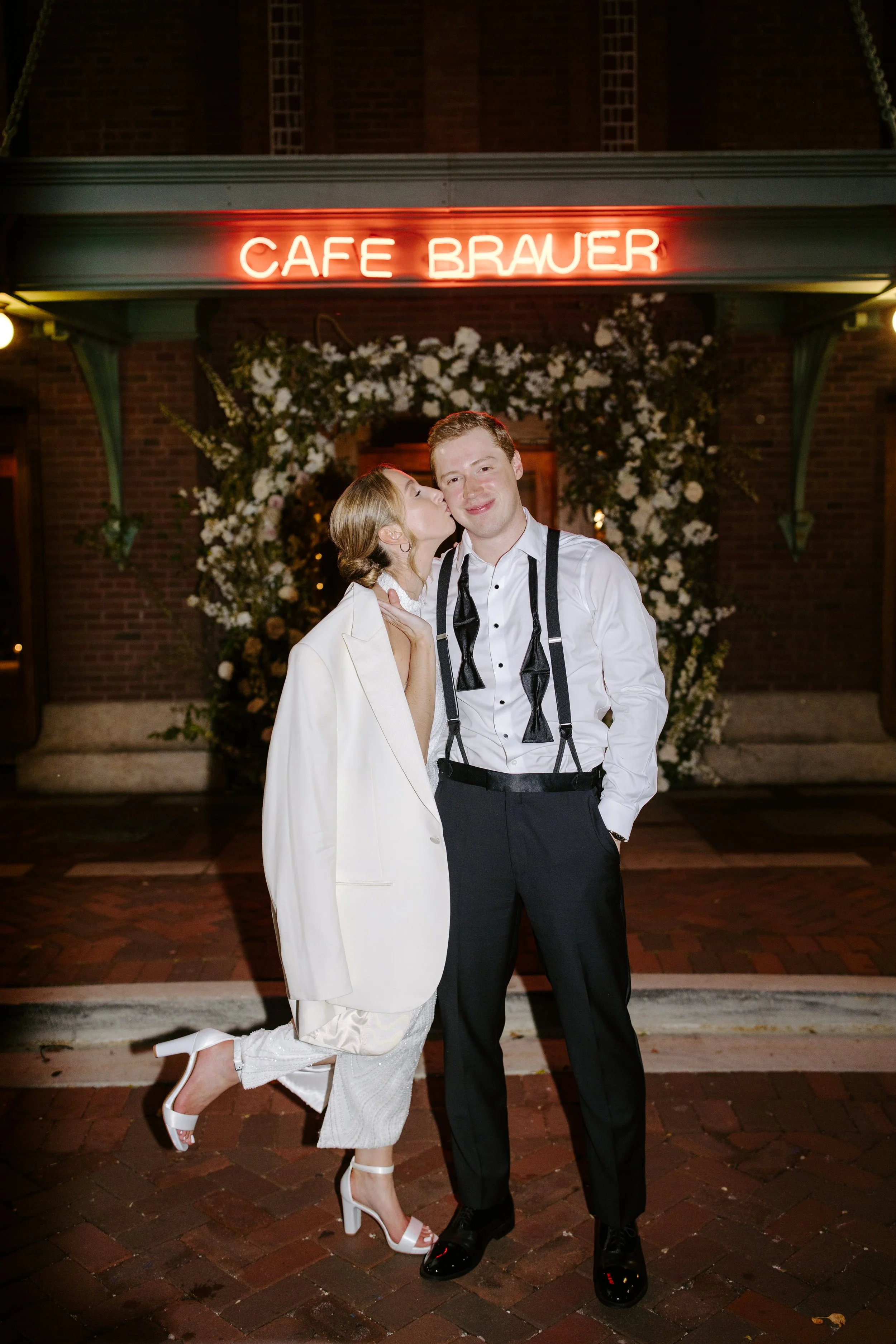 A man and a woman in formal attire standing outside a restaurant with a neon sign reading 'Cafe Brauer'. The woman is dressed in a white blazer and pants, wearing white high heels, and leaning in to kiss the man on the cheek. The man is wearing a whi