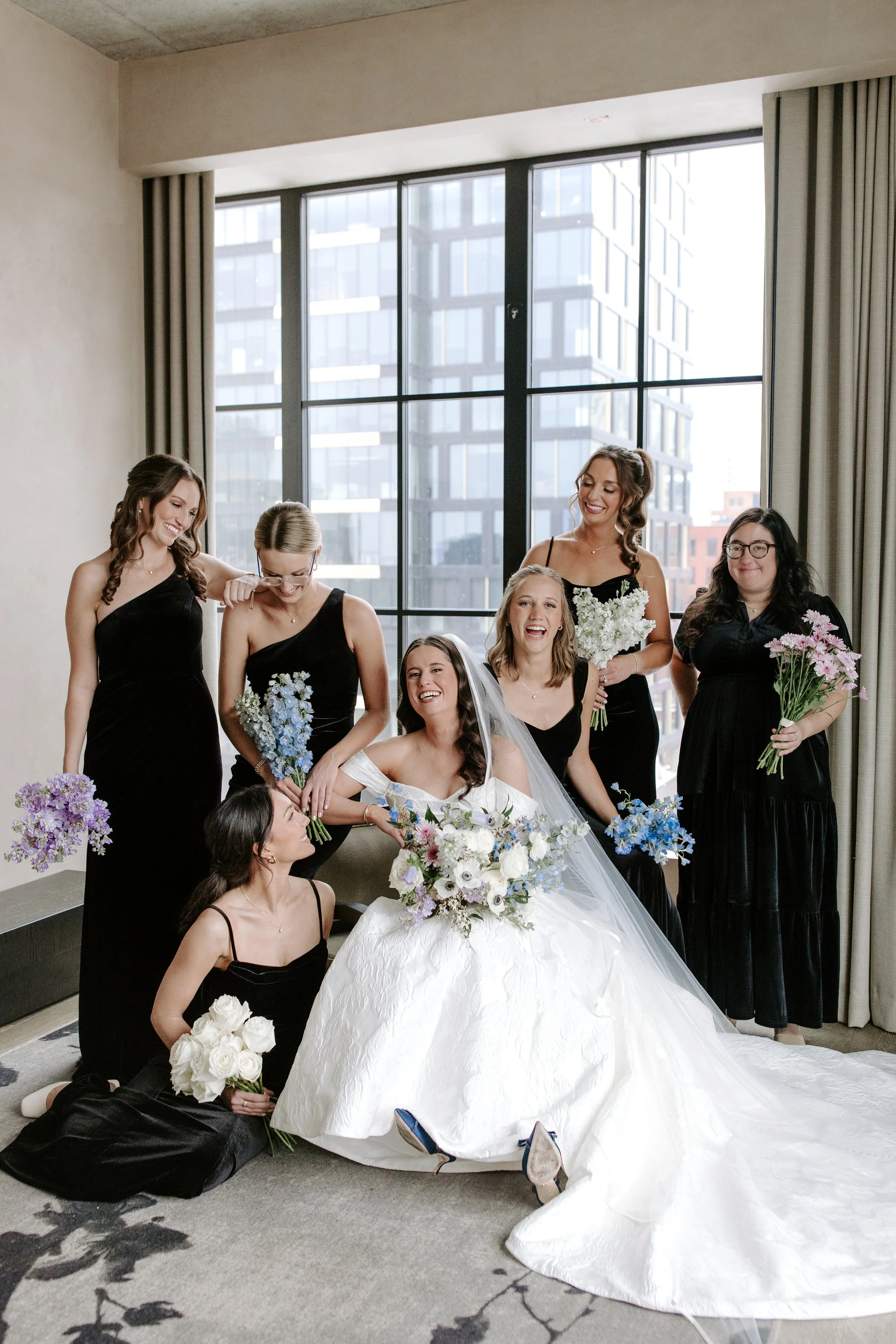Group of women at a wedding, some kneeling and others standing, holding bouquets of flowers, with a large window and city buildings in the background.