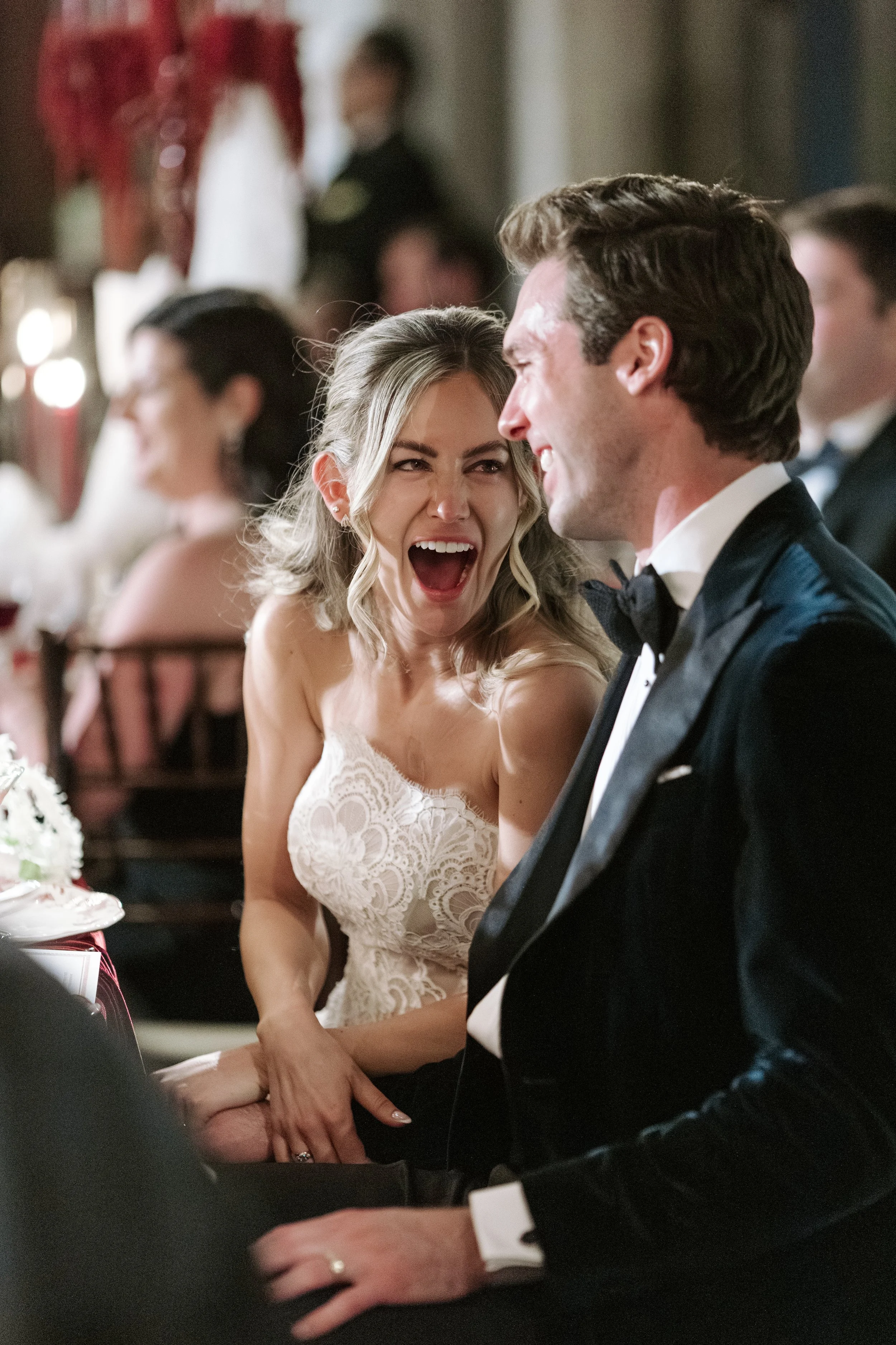 A bride and groom at a wedding reception, sitting closely and smiling joyfully. The bride has wavy blonde hair and is wearing a lace wedding dress, while the groom has wavy brown hair and is dressed in a black tuxedo with a bow tie. They appear to be