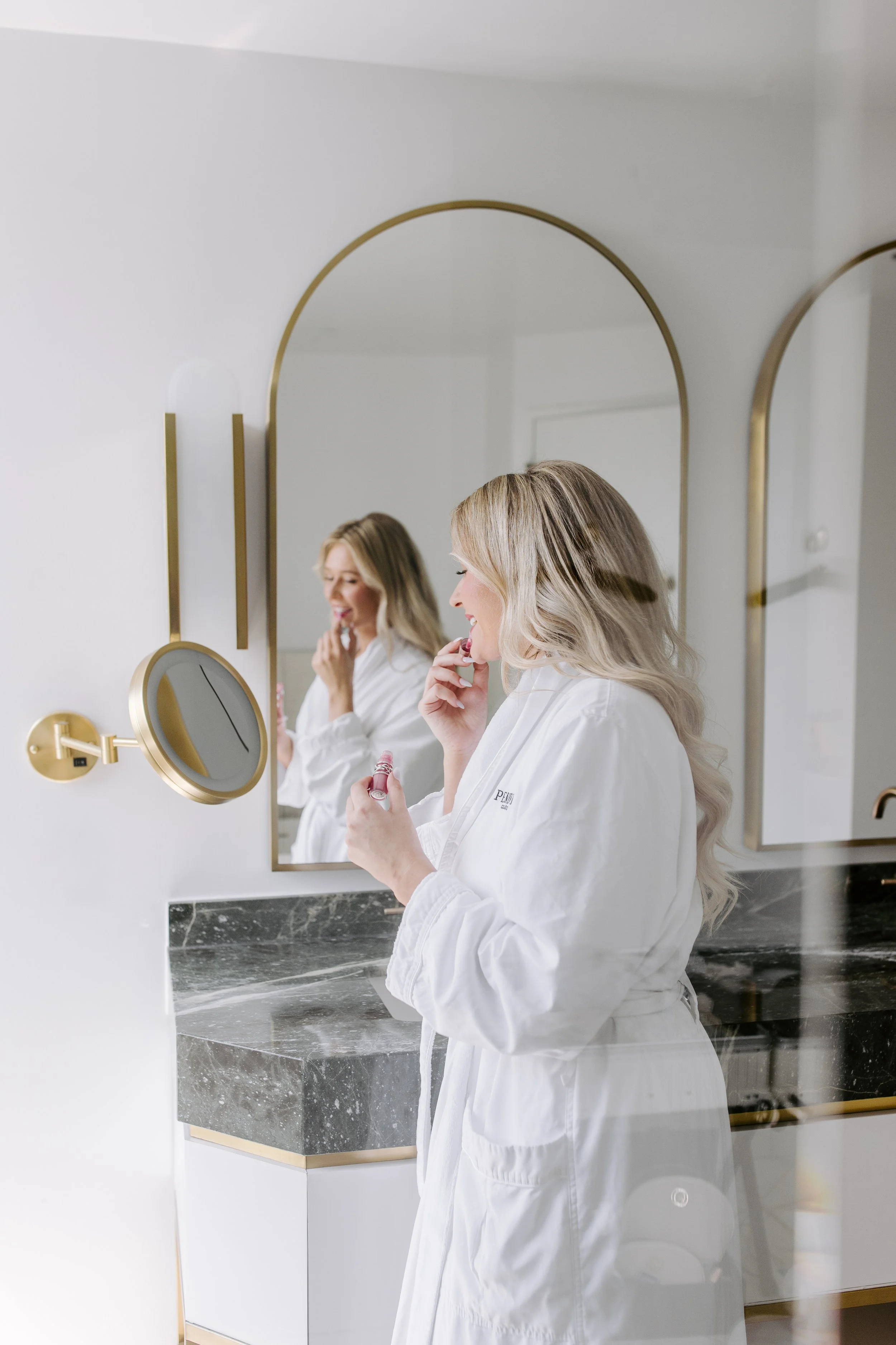 A blonde woman in a white robe is standing at a bathroom sink applying lipstick. She is looking into a mirror with a gold frame, and another mirror of similar style is visible behind her. The bathroom has a marble countertop and a minimalist modern d