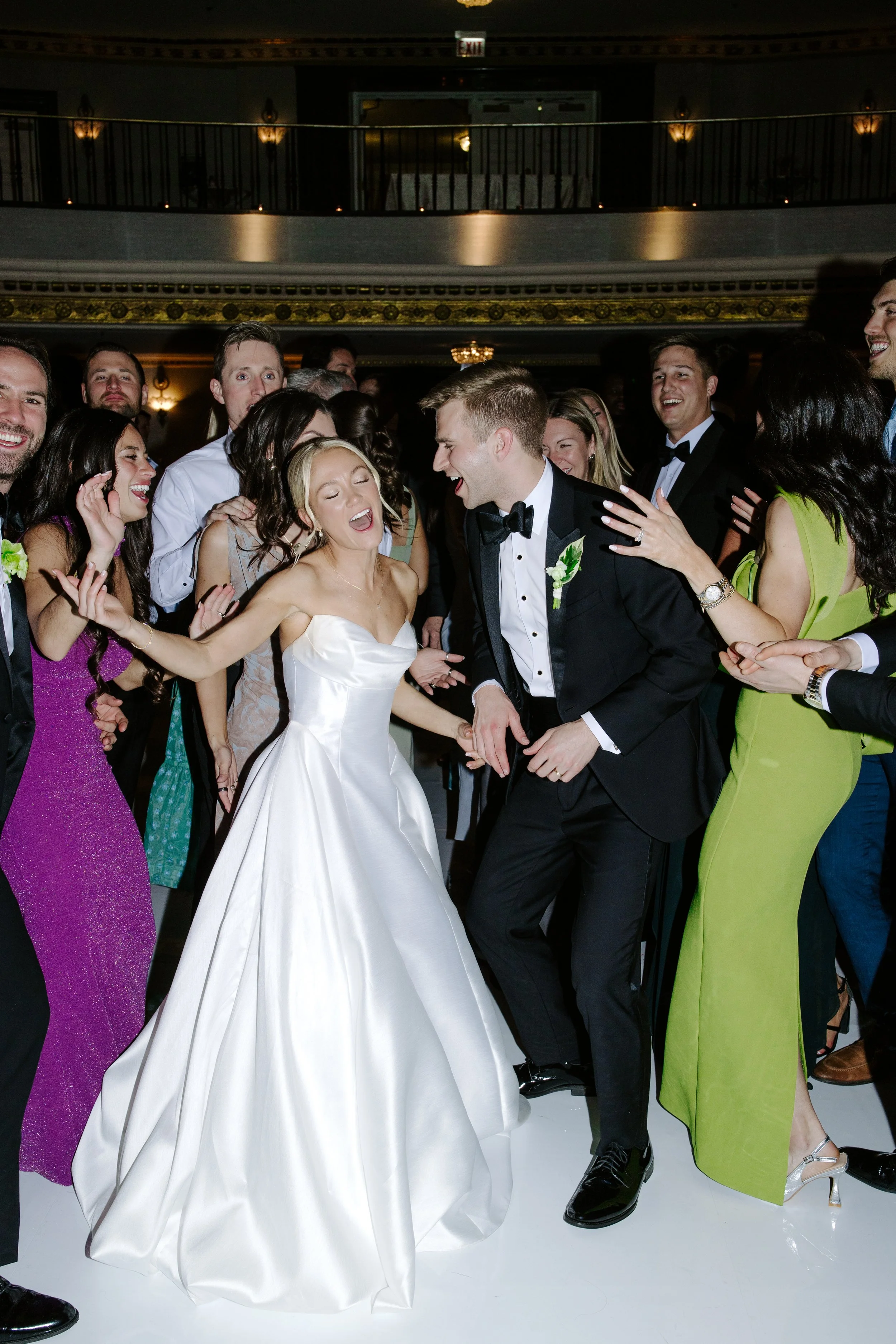 A group of people at a wedding reception dancing and celebrating, with the bride and groom in the center and all dressed in formal attire.